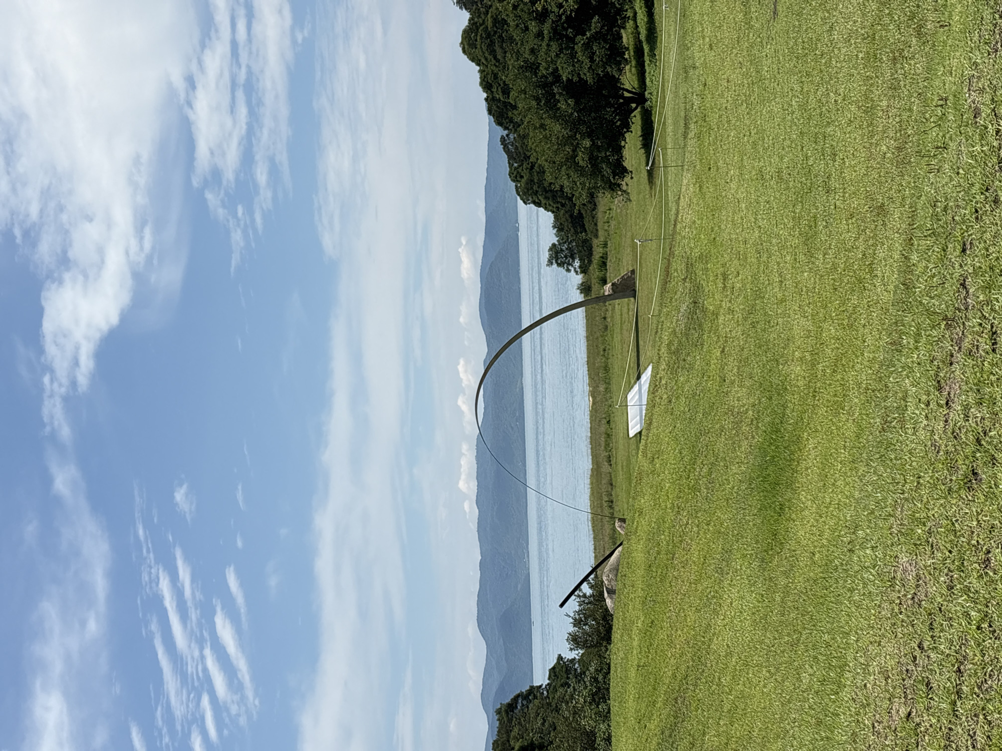 An outdoor landscape scene on the island of Naoshima, Japan, showcases a serene natural environment with a prominent art installation. The foreground is a lush green grassy hill sloping gently towards a body of water. In the mid-ground, a large, curved metallic sculpture dominates the scene, arching gracefully over the landscape. To the left of the sculpture, a large, rough-hewn boulder is visible, with a dark, angled metal pole leaning against it. A white, flat object, possibly part of another installation or signage, rests on the grass in front of the curved sculpture.
The background features a tranquil blue body of water that stretches across the horizon, meeting a chain of distant, hazy blue mountains. The sky above is a vibrant blue, dotted with fluffy white clouds. Dense green trees line the edges of the scene, framing the water and mountains. The overall impression is one of art integrated harmoniously with nature, typical of the artistic landscape found on Naoshima.