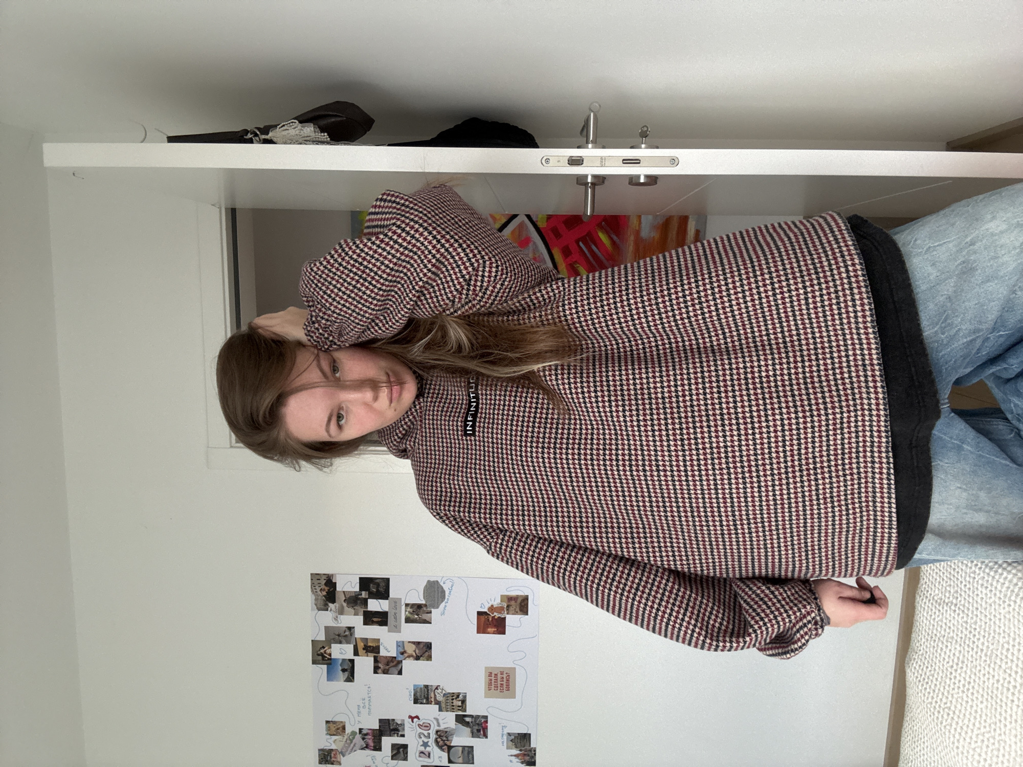 The image depicts an indoor scene within the Vozdovac Urban Municipality, Serbia. A young Caucasian female, estimated to be between 20 and 30 years old, stands centered in the frame, facing the camera directly. She has long, light brown hair, parted on the right, which falls over her shoulders. Her left hand is raised, fingers lightly touching the back of her head, partially affecting her hair. She wears an oversized hooded sweatshirt featuring a red, black, and beige houndstooth pattern. A small black patch with the white text "INFINITUD" is affixed to the chest area of the sweatshirt. A layer of black fabric is visible beneath the hem of the sweatshirt. Her lower body is clad in loose-fitting, light blue denim jeans. Her facial expression is neutral.
The background consists of plain white walls. On the left wall, a white rectangular bulletin board is affixed, covered with numerous small printed photographs, estimated to be between 20 and 30 in number. These photos appear to be personal, depicting various individuals, architecture, and landscapes. Several handwritten notes and drawings in blue marker are present on the board, with visible Cyrillic text including "У меня все получается!", "ciao!", "2+26", "i will love", "скоро на свободу!", and "чтобы вы сделали, если бы не боялись?".
To the right of the subject, a white door is partially ajar. It features a modern, brushed metallic door handle and a lock mechanism, with a key inserted into the keyhole. Behind the open door, a dark-colored handbag with a white beaded or crocheted strap is hanging on a hook. Further into the adjacent space visible through the doorway, a colorful abstract painting with prominent reds, yellows, and blues is discernible. The floor in the lower-left foreground is covered by a light-colored, textured rug or carpet. The scene is illuminated by ambient, possibly natural, light.