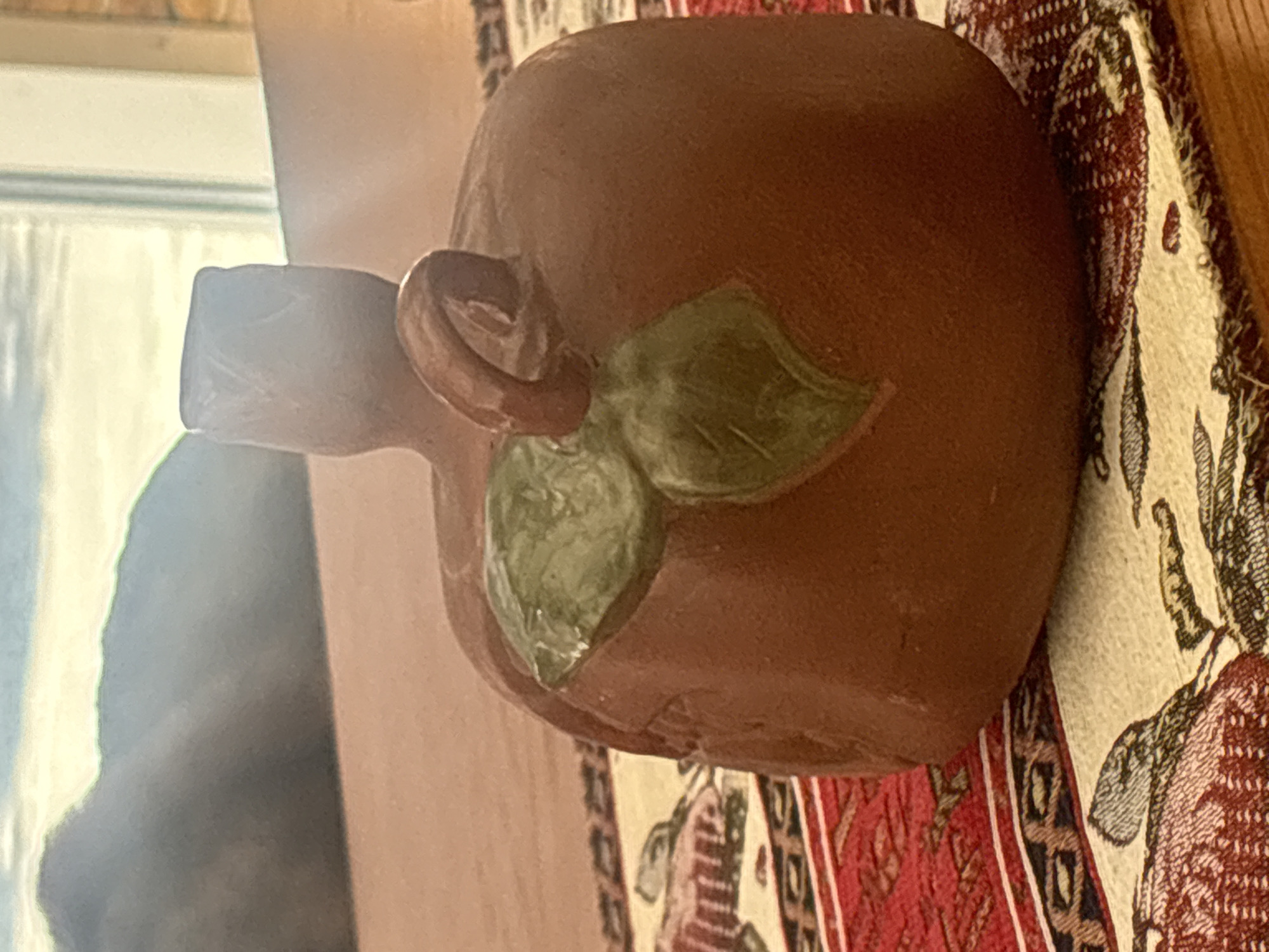 The image displays a close-up of a small, brown, pumpkin-shaped ceramic object with a green leaf and a brown stem. The object is placed on a patterned tablecloth with red and beige sections. In the background, out of focus, is a view through a window showing a landscape with mountains and a hazy sky. The scene appears to be indoors, possibly on a table near a window. There is no information suggesting the location is in the city of Georgiyevsk, Russia.