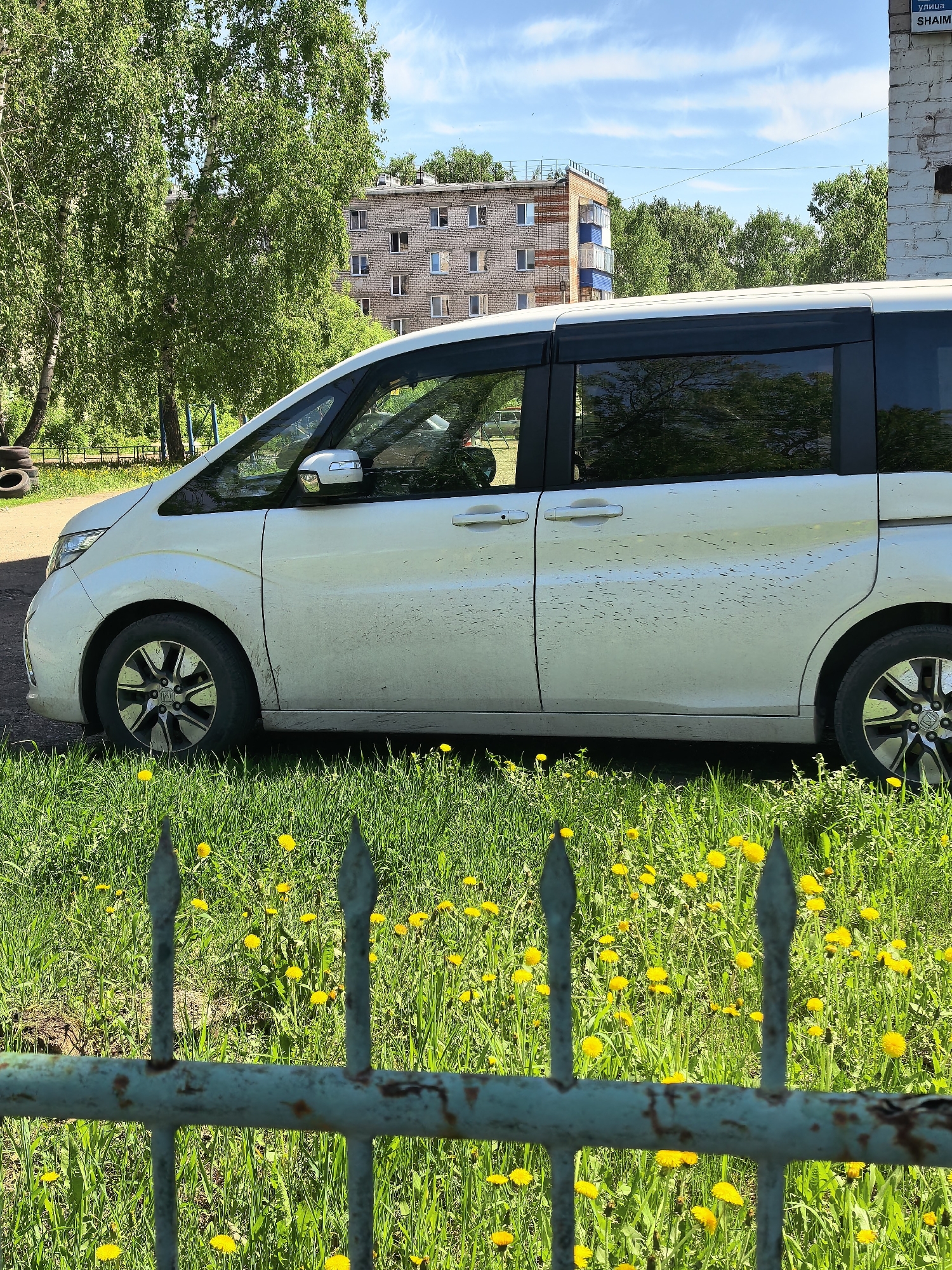 The image depicts a white minivan parked on the edge of a grassy area filled with dandelions. A weathered blue-green metal fence runs in front of the grass and the vehicle, partially obscuring them. The car is visibly dirty.
Behind the car and fence, a residential building is visible among trees under a blue sky with scattered clouds. A building with blue trim stands to the right of the frame, with a blue sign on its side that says УЛИЦА SHAIM (Street SHAIM).
The location is Sterlitamak, Russia, based on the sign mentioning a street named SHAIM, a street located in this city.