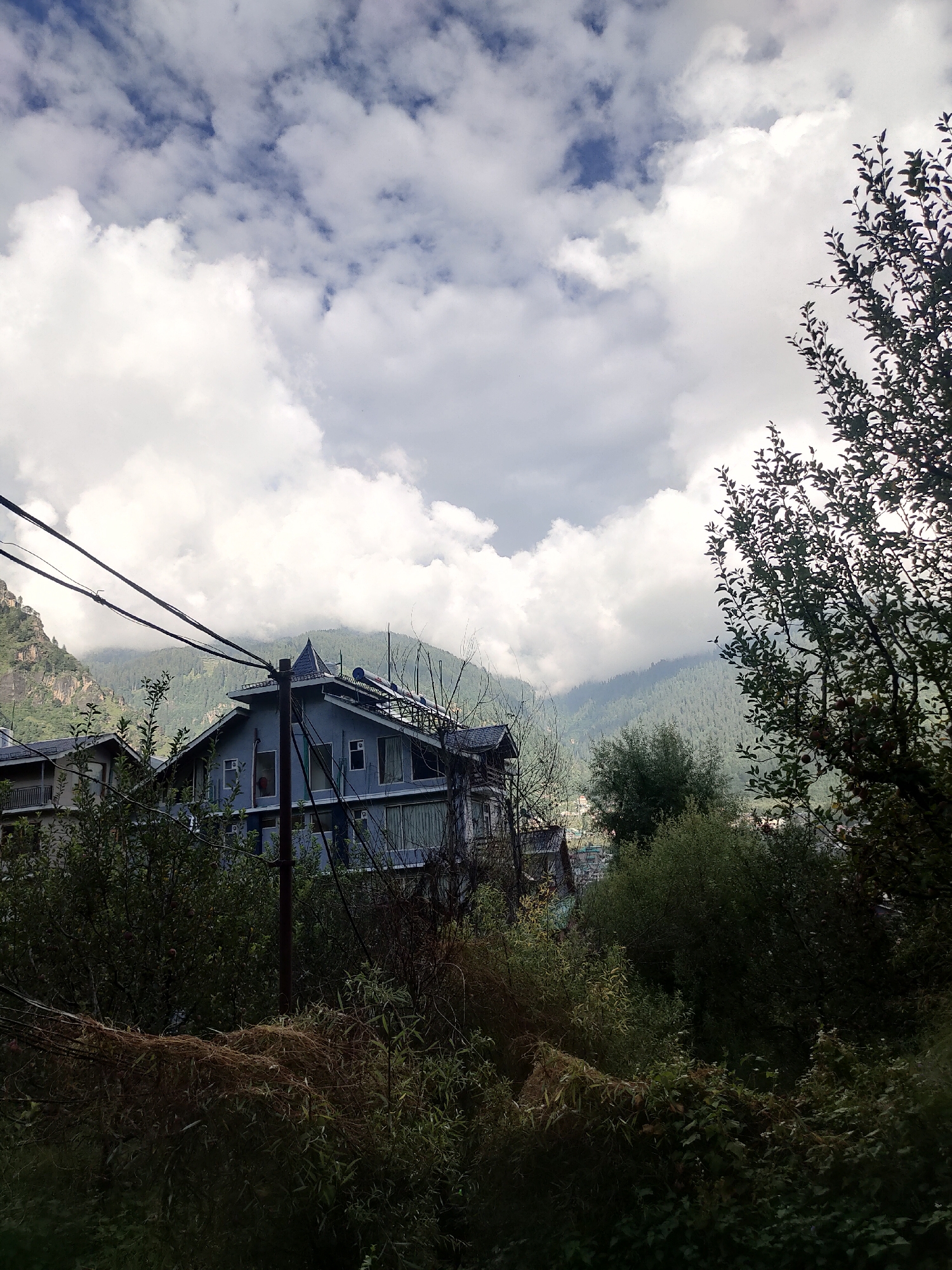 The image depicts a view of a town nestled in a valley with mountains in the background, likely Siumsa, India. In the foreground, there are power lines strung across the frame, supported by a wooden pole. To the left of the pole, a light blue building with several windows and a dark grey, angular roof is visible. To the left of this building, a portion of another building with a lighter facade and balconies can be seen. The middle ground is filled with dense green foliage, with some dry, brown vegetation in the lower portion. The background features the slopes of mountains covered in trees, with some areas obscured by clouds. The sky is a mix of blue patches and large, white, fluffy clouds. No people are visible in the scene.