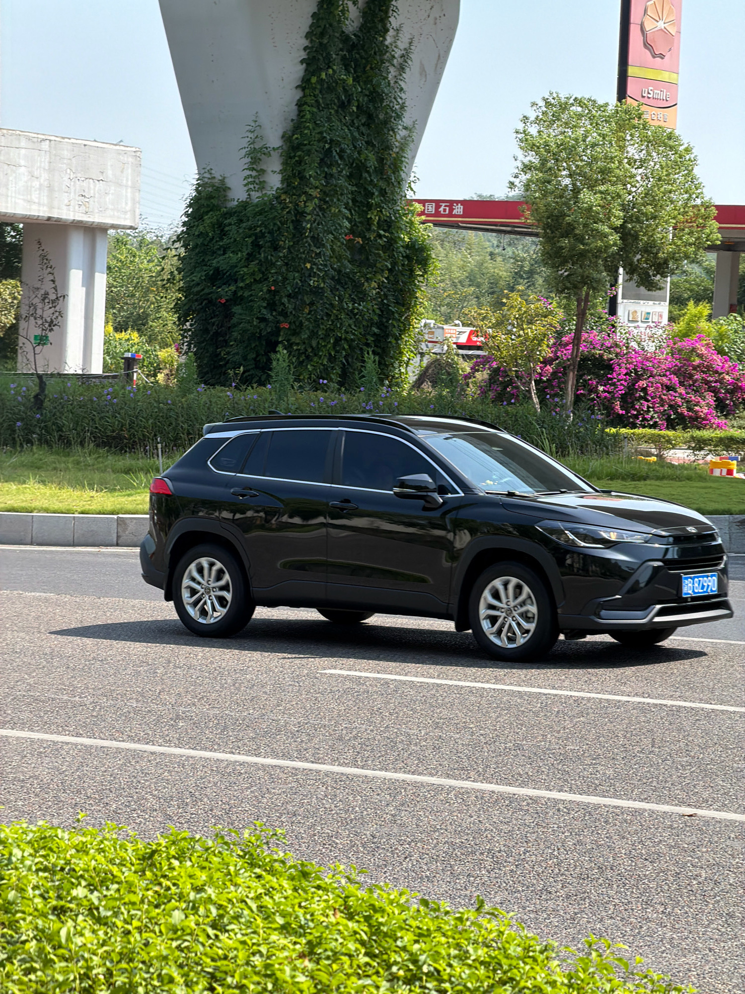 A black SUV, possibly a Toyota Corolla Cross, is driving on a paved road in the city of Jiangjin, China. The vehicle's license plate reads "B 8Z990". In the background, a large concrete structure, likely an overpass, is covered in lush green ivy. To the right of the overpass, a gas station with a red canopy is visible, featuring a sign with the text "uSmile" and Chinese characters. Several trees and vibrant pink and purple flowers are also present in the scene. The road is marked with white lines, and a strip of green bushes lines the foreground of the image. The sky is clear and blue.