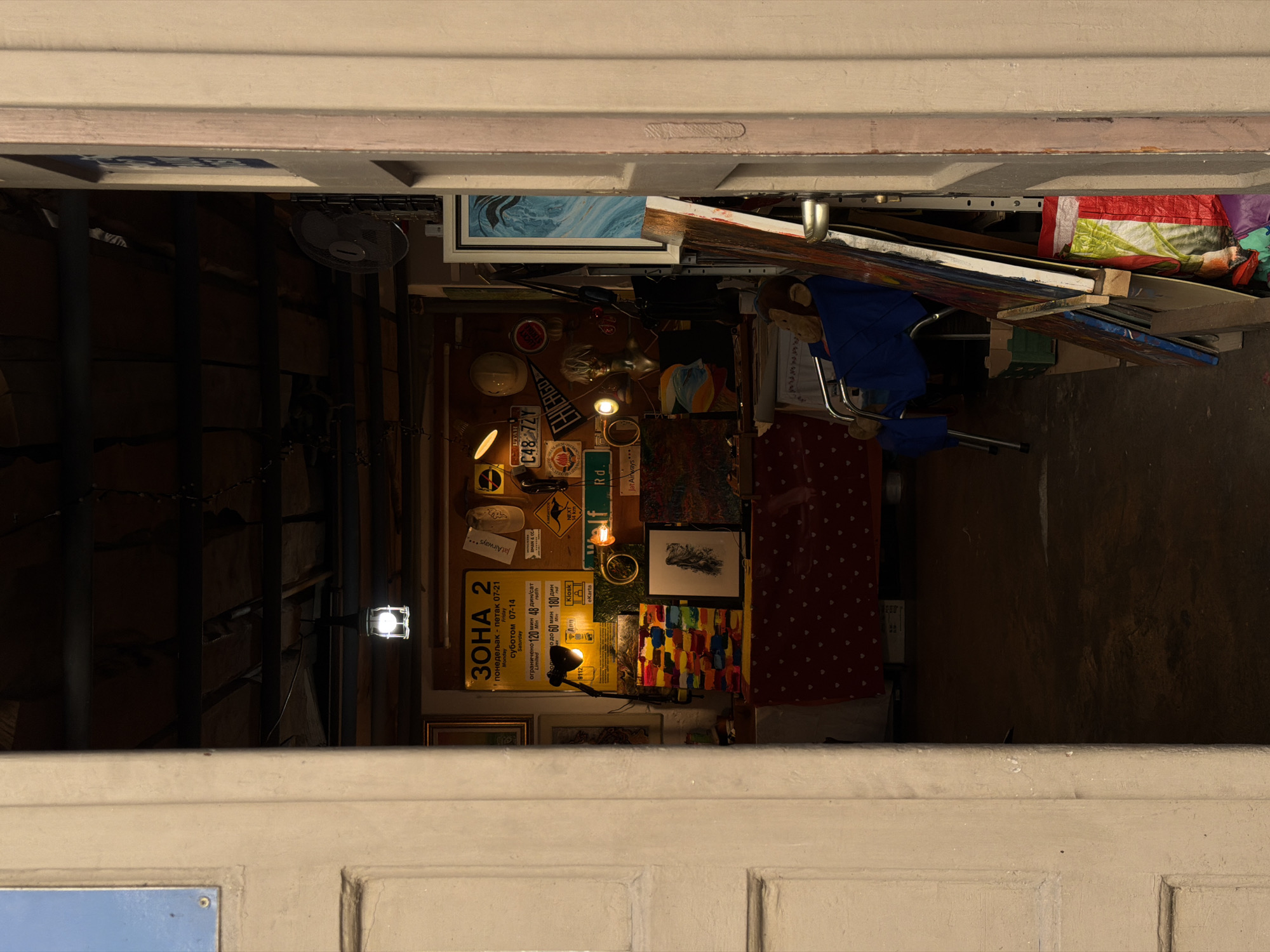 View from inside a building in Belgrade, Serbia, through a doorway or window. The interior is cluttered with items, including artwork, a fan, license plates (one reads "C48 72Y," another has "CHICAGO"), signs in Serbian Cyrillic, and a blue chair. The location may be a shop or workshop. One sign includes the word "eKarta."
