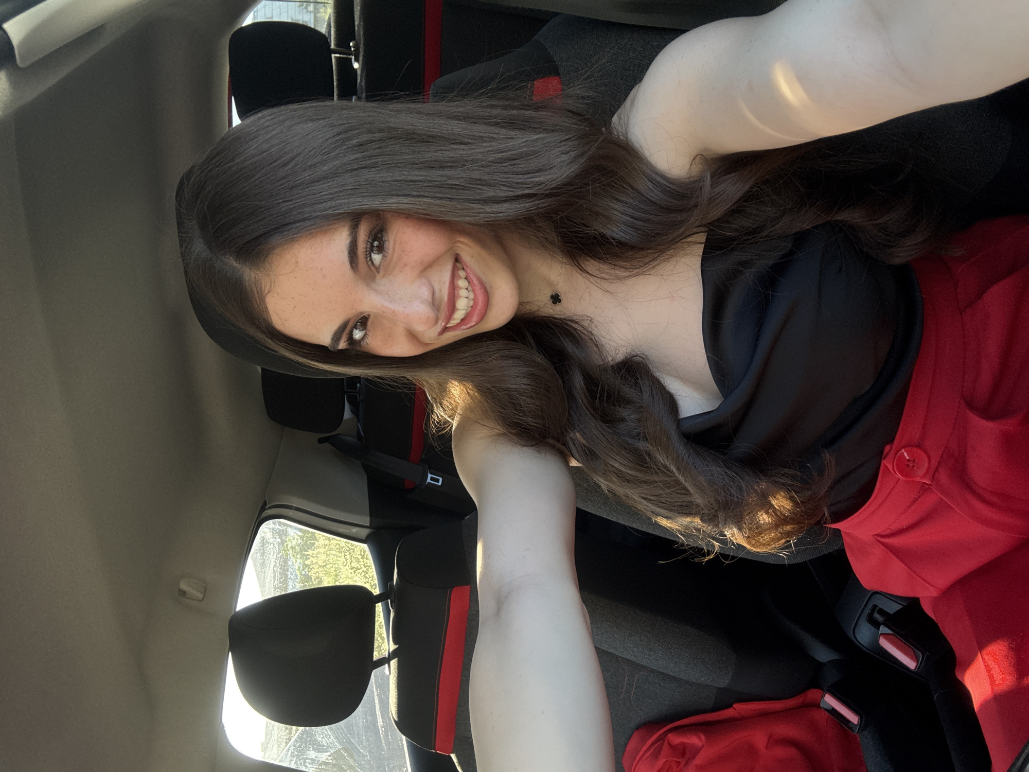 A young woman with long brown hair and fair skin is taking a selfie inside a car. She is wearing a black top and red pants. The interior of the car is visible, including the seats and seatbelts. The background shows a view out the car window, appearing to be a street with vegetation and possibly buildings in the distance. The location in the city of Plovdiv, Bulgaria cannot be determined from the image.