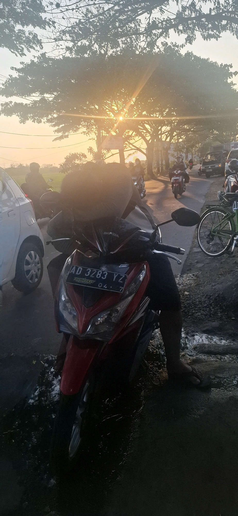 In Desa Pandeyan, Indonesia, a red motorcycle is parked on a gravelly surface with puddles reflecting the hazy, golden light of sunset filtering through large trees overhead. A person wearing dark shorts sits on the motorcycle, their legs visible. The motorcycle has a license plate reading "AD 3283 YZ". In the background, other vehicles, including a white car, a dark van, and several other motorcycles, are visible on a road lined with trees. The sky is a bright yellow and orange, indicating a sunset.