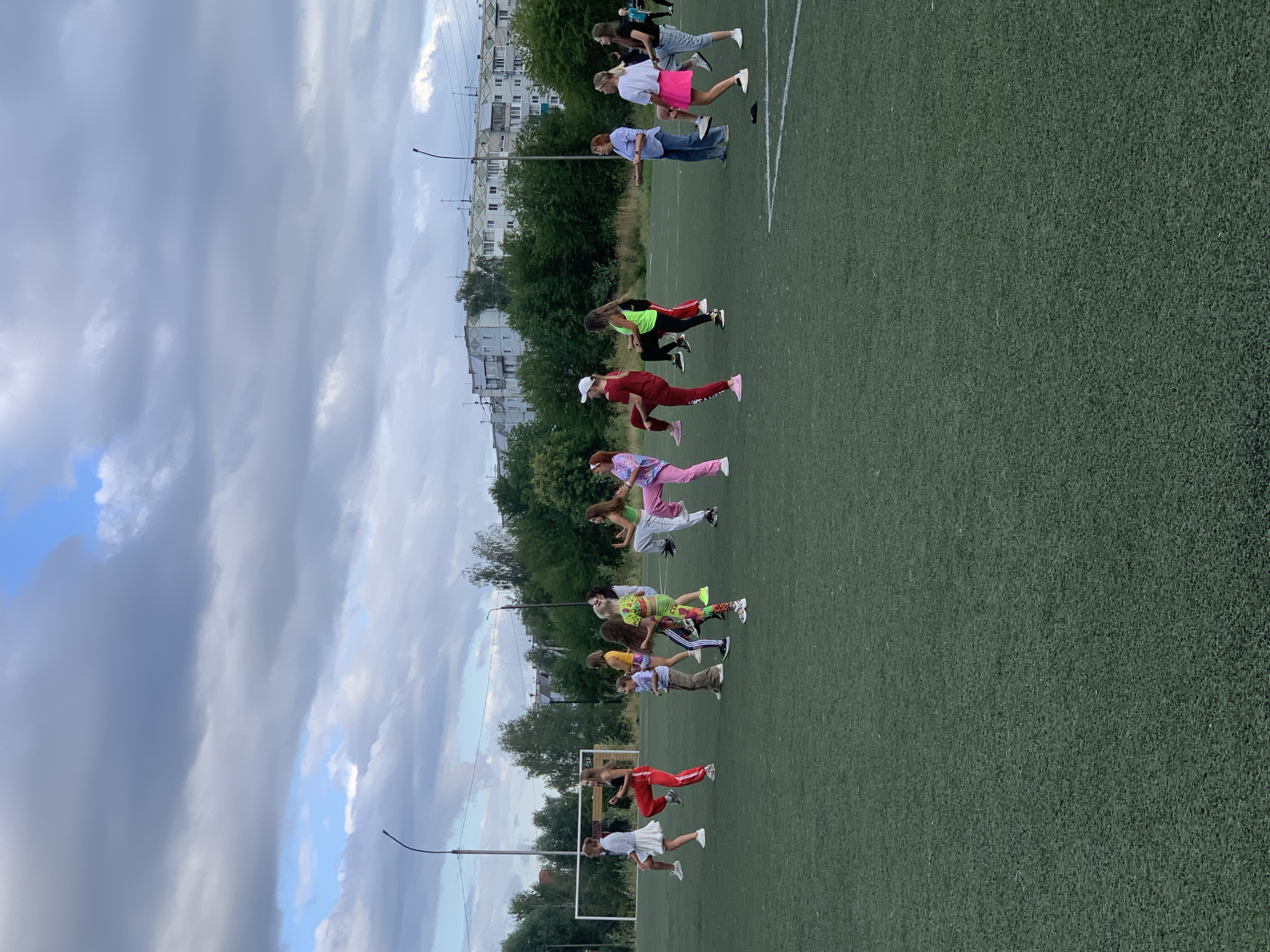 A group of people are engaged in a dance class on a green artificial turf field, likely a stadium or sports complex. The group consists of individuals of various ages, with a majority appearing to be young women and girls. They are dressed in athletic or casual wear, with some wearing vibrant colored outfits. The individuals are spread out across the field, performing synchronized movements, indicating a choreographed activity. In the background, there is a soccer goal, trees, and residential buildings typical of a city environment. The sky is cloudy, suggesting an overcast day. The presence of the sports facilities and urban background points to a location within a city. Given the context provided, this scene is located in Prokopyevsk, Russia.