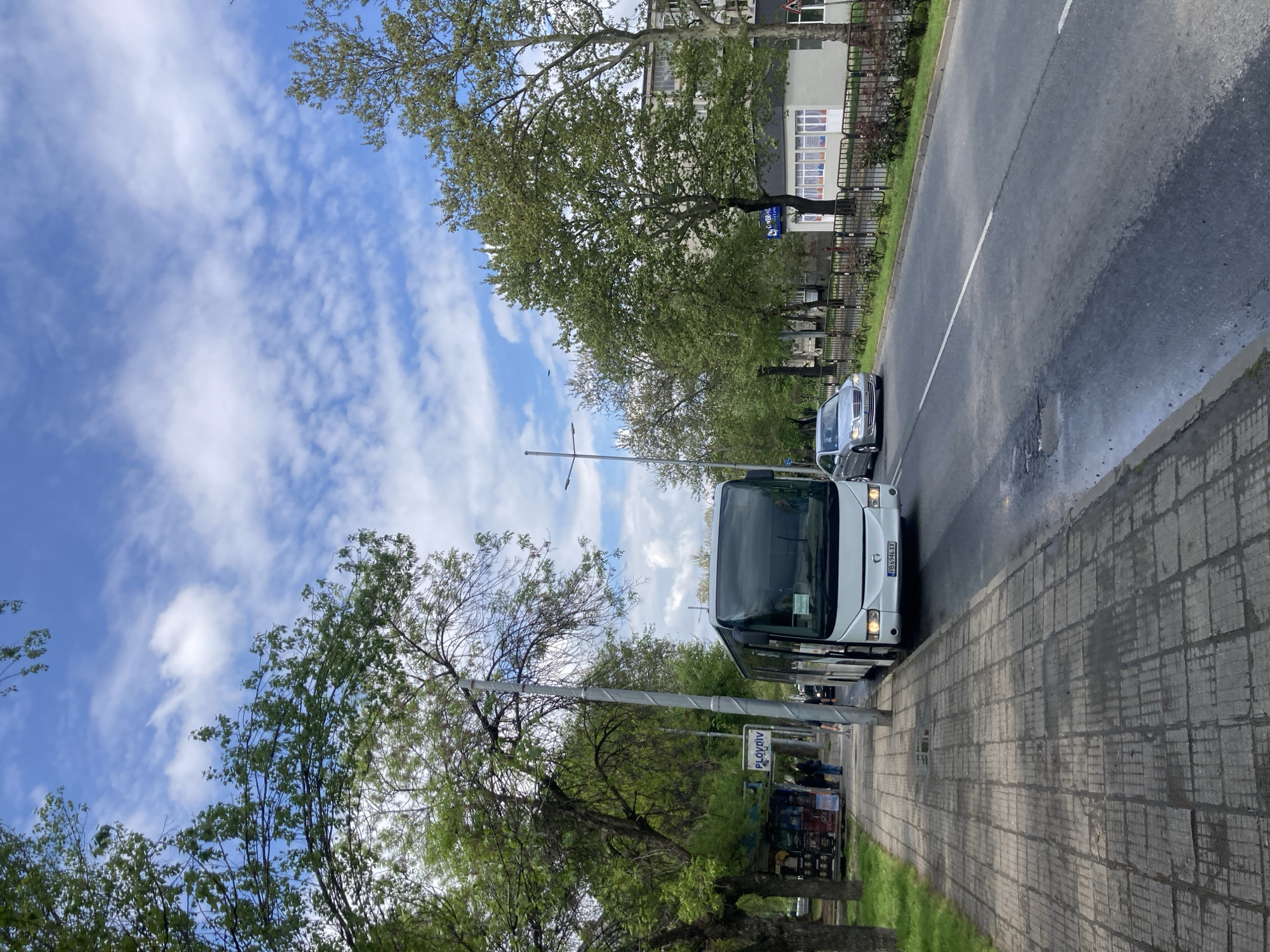 The image shows a city street with a white bus parked on the right side of the road next to a sidewalk. A white car is seen in the distance on the road. The sidewalk is paved with gray tiles. A sign with "PLOVDIV" is visible. Trees line the sidewalk. The sky is blue with scattered white clouds. A street light is on the left side of the bus. The license plate number of the bus is PB6946XK. The location is in Plovdiv.