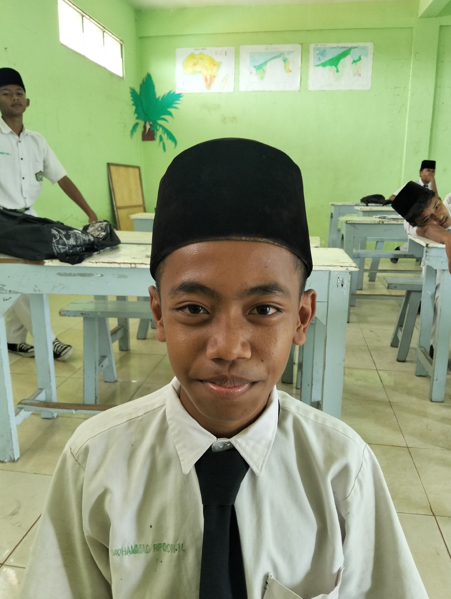 A young male, wearing a white collared shirt with a black tie and a black peci (a cap worn by Muslim men), is pictured in a classroom in Kranji, Indonesia. The student is positioned centrally in the foreground, facing the camera with a slight smile. Behind him, other students and school desks are visible. The classroom walls are painted green, and educational posters, including maps, are displayed. Natural light streams in from windows at the top of the image. The overall impression is of a typical school day setting.