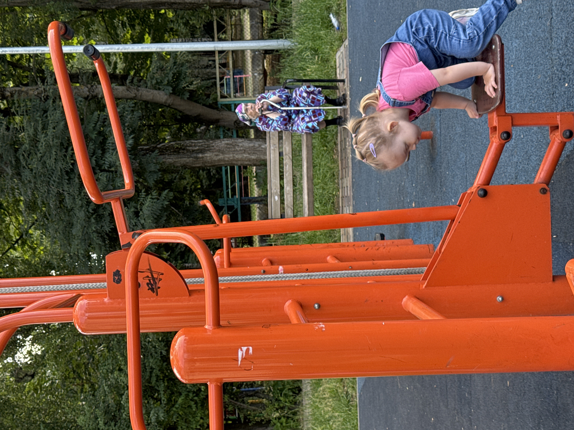In Viazniky, Russia, on an outdoor fitness equipment area, a young Caucasian girl with blonde pigtails and a pink shirt and denim overalls is crouched on an orange exercise machine. Her hands are on the machine's seat, and she appears to be climbing onto it. In the background, an elderly Caucasian woman with white hair is seated on a wooden bench, holding a cane and wearing a brightly patterned robe and headscarf. The fitness equipment is made of orange metal tubes. Behind the bench, trees and some fencing are visible, suggesting a park or public space. The ground is covered with blue rubber matting.