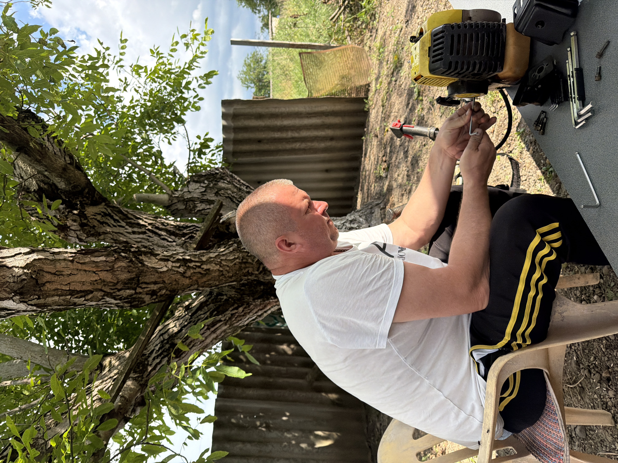 A man in a white t-shirt and black athletic pants with yellow stripes is sitting on a plastic chair and working on a yellow and black gas-powered string trimmer. He is holding a hex wrench and appears to be tightening or loosening a bolt on the trimmer's engine. Various tools, including other hex wrenches and bolts, are laid out on a gray table in front of him. The scene is outdoors, with a large tree providing shade. In the background, there is a corrugated metal fence, some dry vegetation, and a cloudy sky. The ground is covered in dirt and small stones. The man is fair-skinned with a shaved head and is looking down at his work. The overall impression is of a casual outdoor repair or maintenance activity.