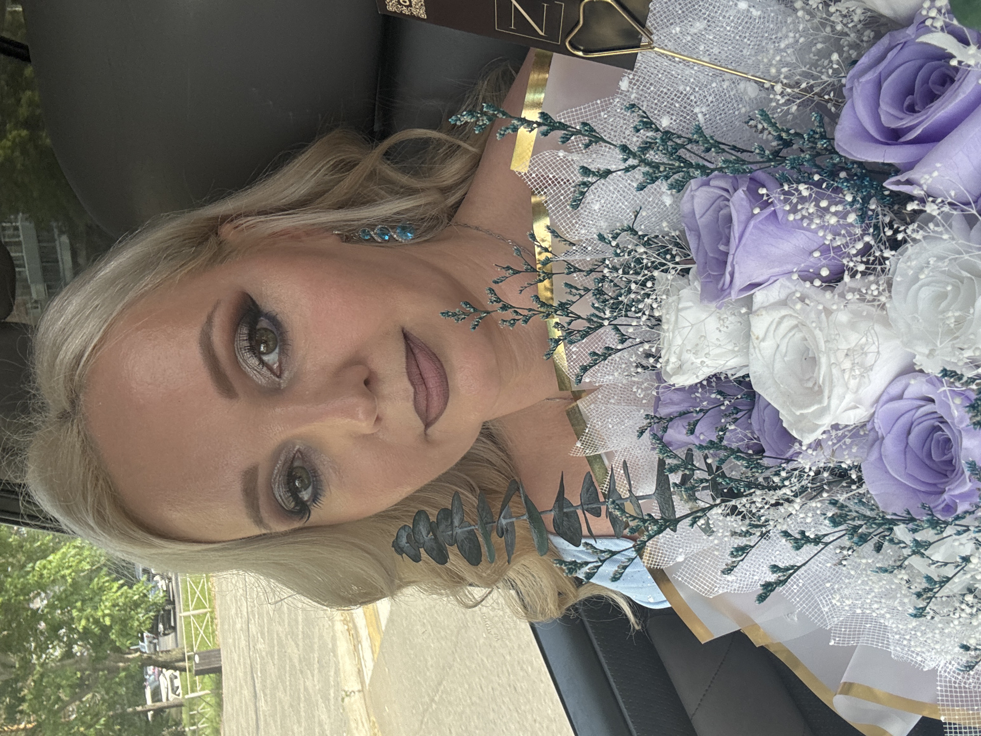 A blonde woman is seated in the passenger seat of a vehicle, holding a bouquet of preserved roses and filler flowers. The bouquet is primarily composed of white and lavender roses, accented with delicate white and teal foliage. The woman is looking towards the camera, her makeup applied with dramatic winged eyeliner and muted lipstick. She is wearing a light blue off-the-shoulder top and dangling turquoise earrings. The background shows a blurred view of trees, parked cars, and a building, consistent with an urban environment in Komsomolsk-on-Amur, Russia. The interior of the car, including the dark headrest, is also visible. The image appears to be a selfie taken by the woman.