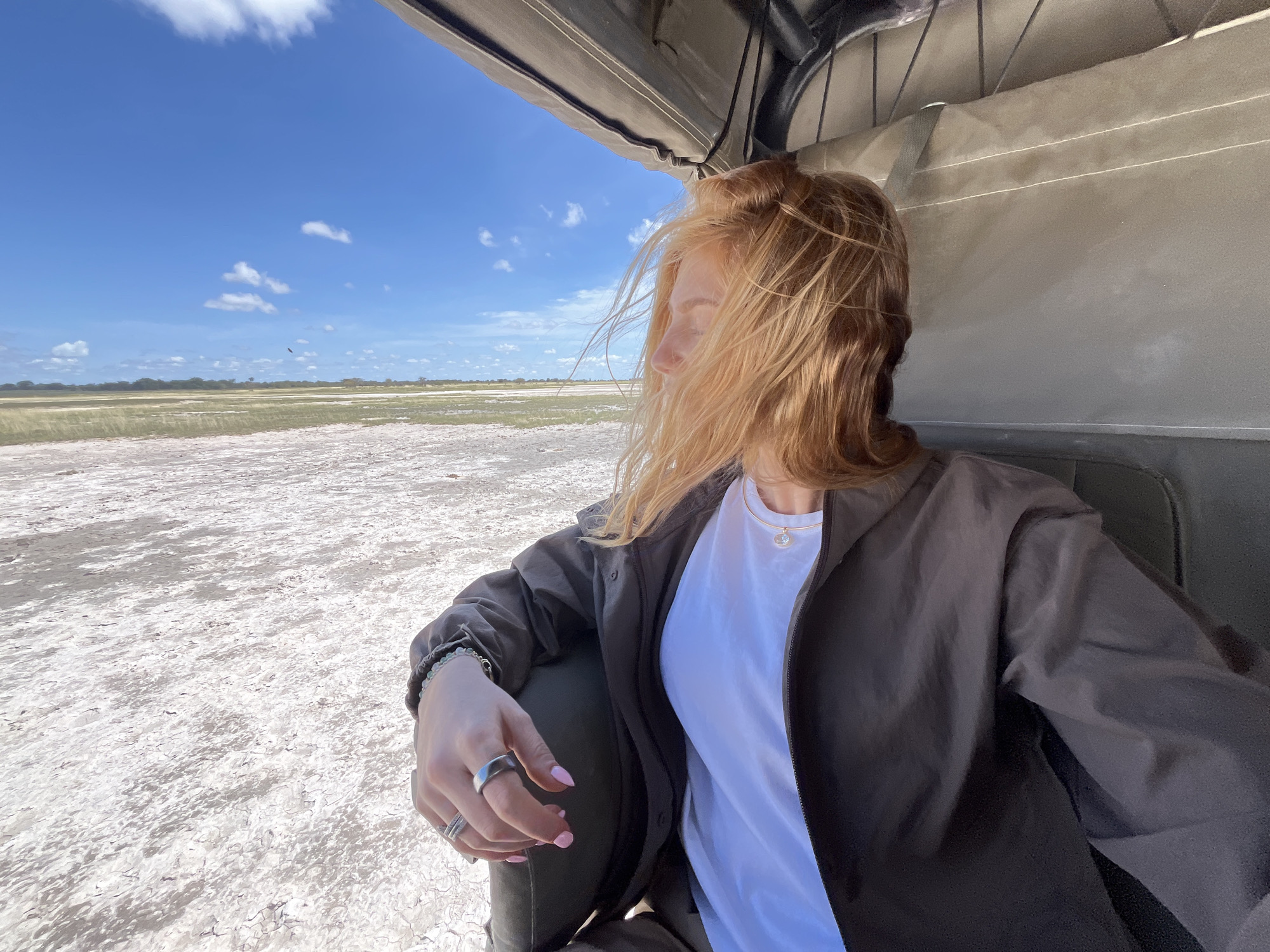 A young, fair-skinned person with ginger hair, wearing a white t-shirt and a dark brown jacket, is seated inside an open-topped vehicle with a light khaki interior. The person is looking out toward a vast, flat, dry landscape dotted with sparse vegetation under a bright blue sky with scattered white clouds. The surface of the landscape appears cracked and white, suggesting a dry, arid environment. The scene is reminiscent of the Makgadikgadi Pans in Botswana, near Gucta. The person is wearing rings and a bracelet. The vehicle is providing shelter from the sun with its canvas roof. The overall impression is of a safari trip or exploration in a remote, arid location.