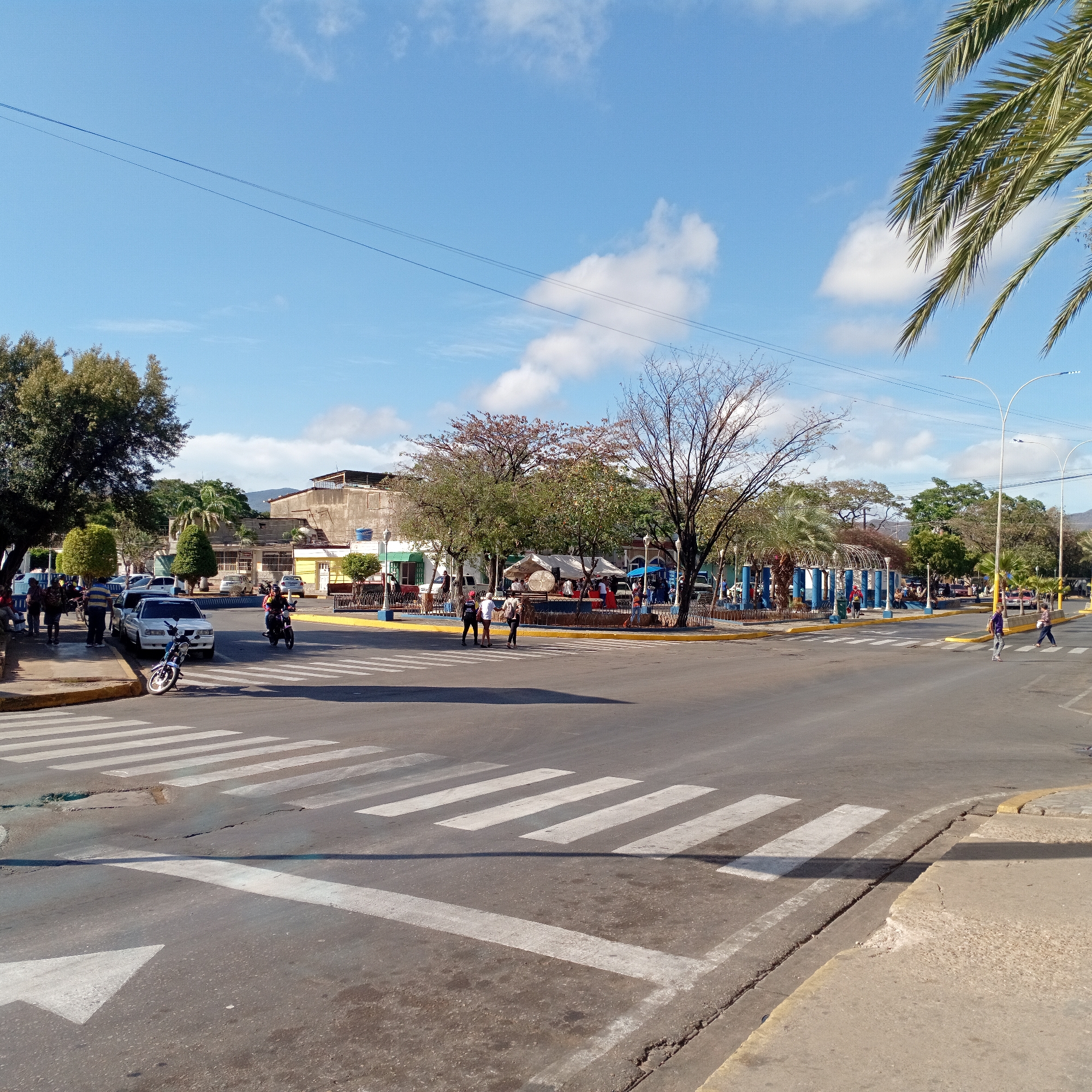 The image depicts an intersection in a city. The sky is blue with scattered clouds. In the foreground, there is an asphalt road with white crosswalk markings and a white arrow indicating a direction. The road surface shows signs of wear and tear.
To the left, there is a sidewalk with pedestrians standing and walking. Several parked cars are visible along the sidewalk. One motorbike is leaning over slightly.
In the middle ground, a central area of trees and a seating area exists; people are present around the perimeter of the park.
In the background, buildings of varying sizes are seen, partially obscured by trees. Overhead power lines stretch across the sky. A palm tree is present in the top right corner.