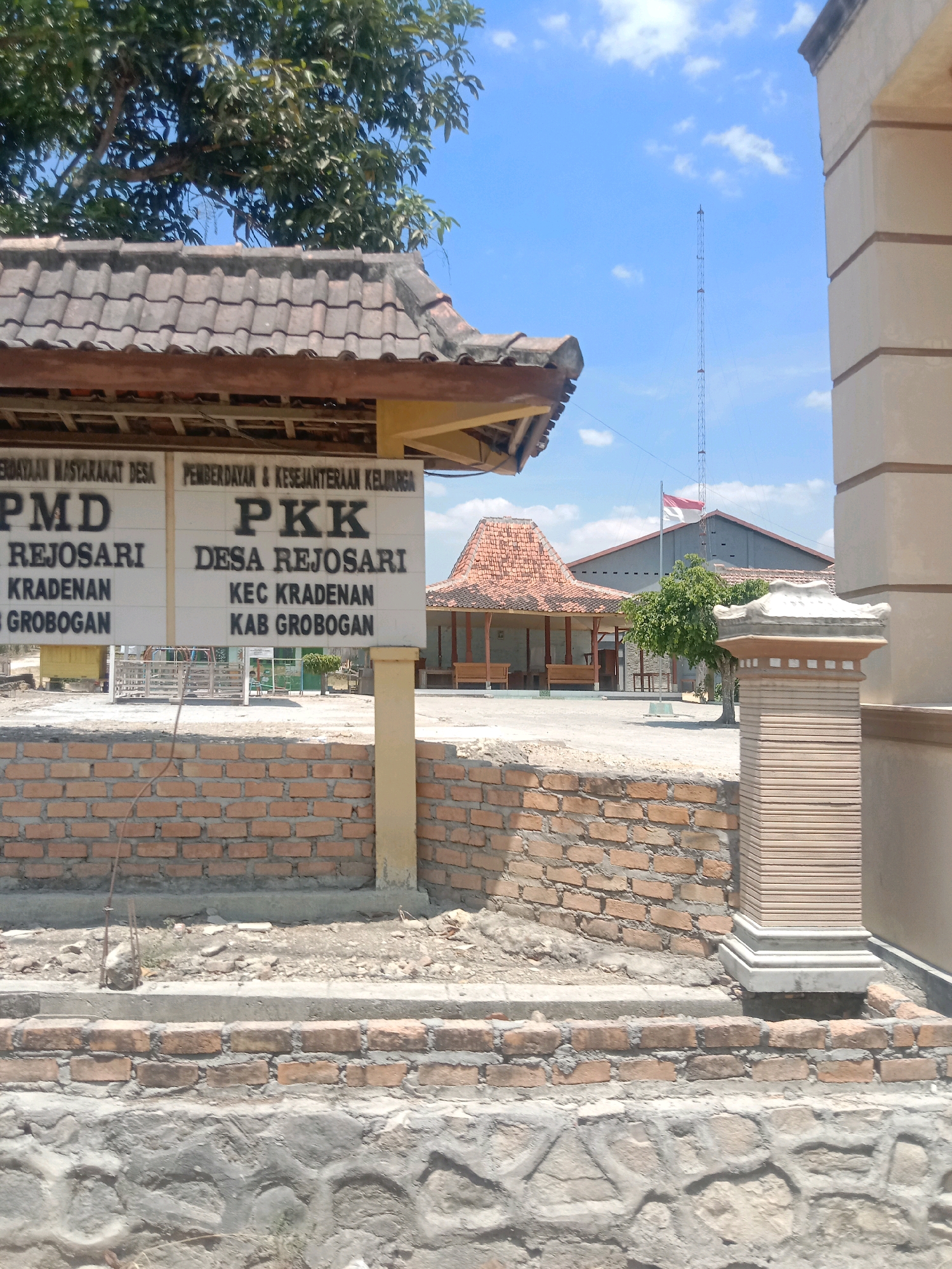 The image captures a view of the Rejosari village hall in Grobogan, Indonesia, under a clear blue sky with scattered clouds. In the foreground, a brick wall and a decorative pillar are visible. Behind the wall, a sign displays text in Indonesian, indicating "PMD REJOSARI," "KRADENAN," and "KAB GROBOGAN" on the left, and "PKK DESA REJOSARI," "KEC KRADENAN," and "KAB GROBOGAN" on the right. These indicate the name of the village, sub-district, and district. The background features traditional Indonesian architecture with red tiled roofs, including a main building and smaller structures, suggesting a public gathering space or administrative center. A tall communication tower and the Indonesian flag, a red and white bicolor, are also prominent elements in the scene, symbolizing national presence. The overall environment appears to be a sunny, outdoor setting in a rural or semi-urban area of Indonesia.