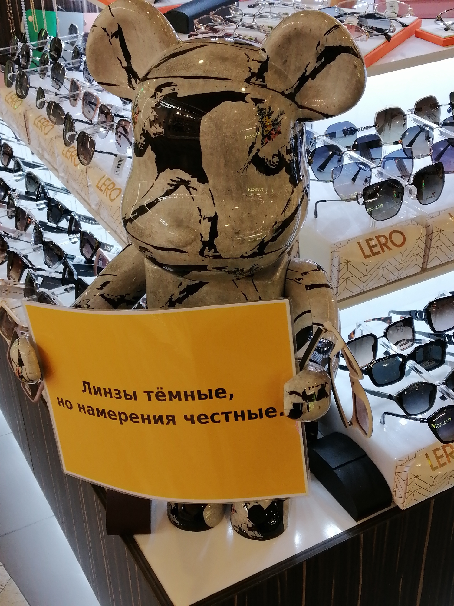 This media file showcases a retail display, likely in an optical store in Rostov-on-Don, Russia. The focal point is a large, stylized bear-shaped figure, decorated with a black and white abstract pattern that includes floral elements. The figure is holding a bright yellow sign with black text.
The sign reads: "Линзы тёмные, но намерения честные." This translates to "The lenses are dark, but the intentions are honest." This playful message suggests a humorous approach to marketing eyewear, perhaps alluding to sunglasses or prescription glasses with tinted lenses.
Behind the bear figure, numerous pairs of sunglasses are displayed on racks. These glasses vary in style and color, with many featuring dark lenses. Some of the sunglasses have brand names visible, such as "LERO". The overall setting is brightly lit, indicative of an indoor retail environment. The presence of multiple displays and the organized arrangement of products suggest a commercial space.
The time of day and weather conditions are not discernible from the image. The overall mood is lighthearted, conveyed by the whimsical bear figure and the humorous text on the sign. The location is specified as Rostov-on-Don, Russia.