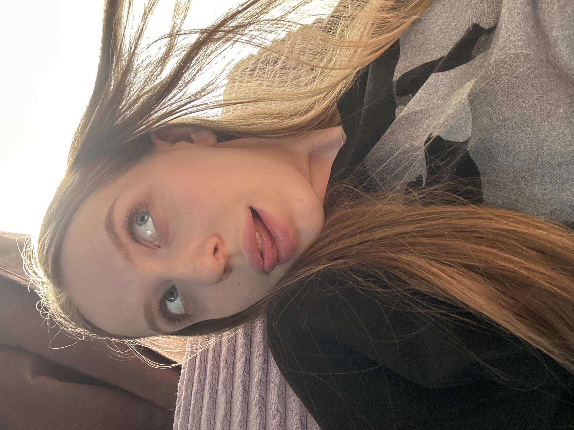A young woman with light blonde and brown hair, blue eyes, and fair skin is shown in an angled selfie. Her hair is partially up, and she is wearing a black long-sleeved shirt with a gray patterned overlay. The background includes a light-colored corduroy surface and bright, diffused light. The image's location in Chaikovsky, Russia, cannot be verified.