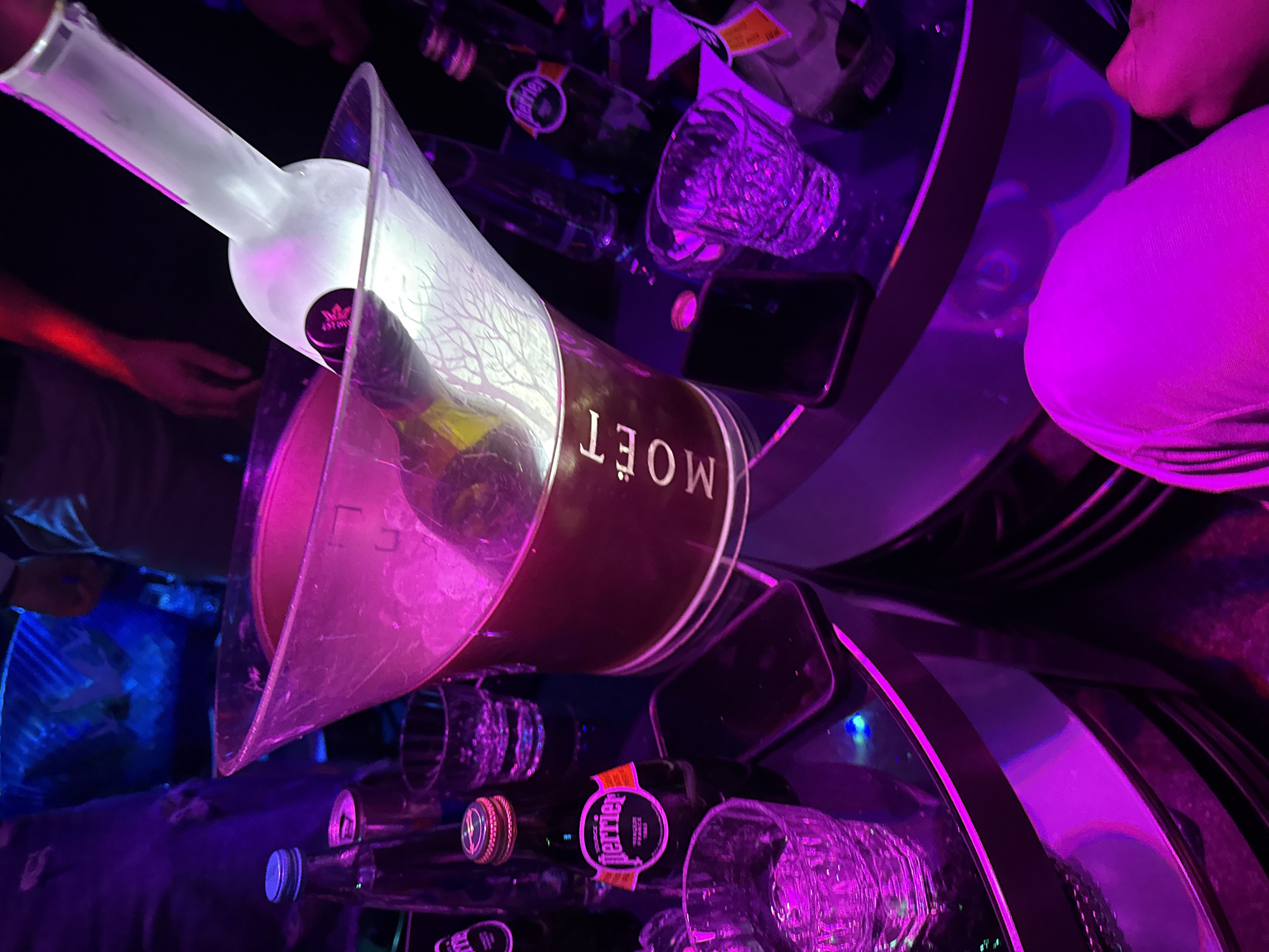 The image depicts a dimly lit scene, likely indoors, possibly a nightclub or bar. There's a Moët champagne bucket containing a frosted bottle of Grey Goose vodka. The table is covered in bottles of Perrier sparkling water, glasses, and a cell phone. The lighting is heavily saturated with blue and magenta hues. A person's arm is visible in the background. It is difficult to ascertain the precise location in Plovdiv, Bulgaria, from this image alone.