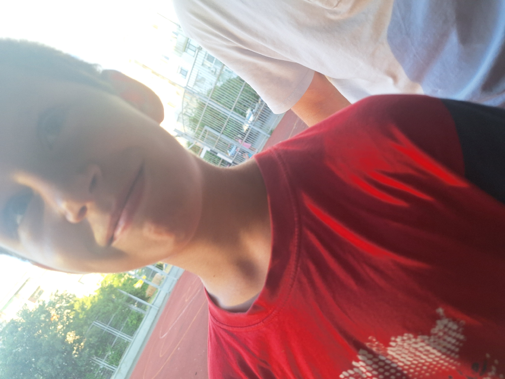 The image shows a close-up, slightly blurred selfie taken outdoors during the day. The foreground is dominated by the upper body of a young person wearing a red t-shirt with a white design. To the right, another person is visible, wearing a white t-shirt. The background features a red sports court with white markings, a metal fence, trees, and buildings. The lighting suggests it is sunny, with the sun possibly behind the camera, causing some overexposure. The scene appears to be a sports facility or playground, possibly within a residential area in Yambol, Bulgaria, given the context provided.