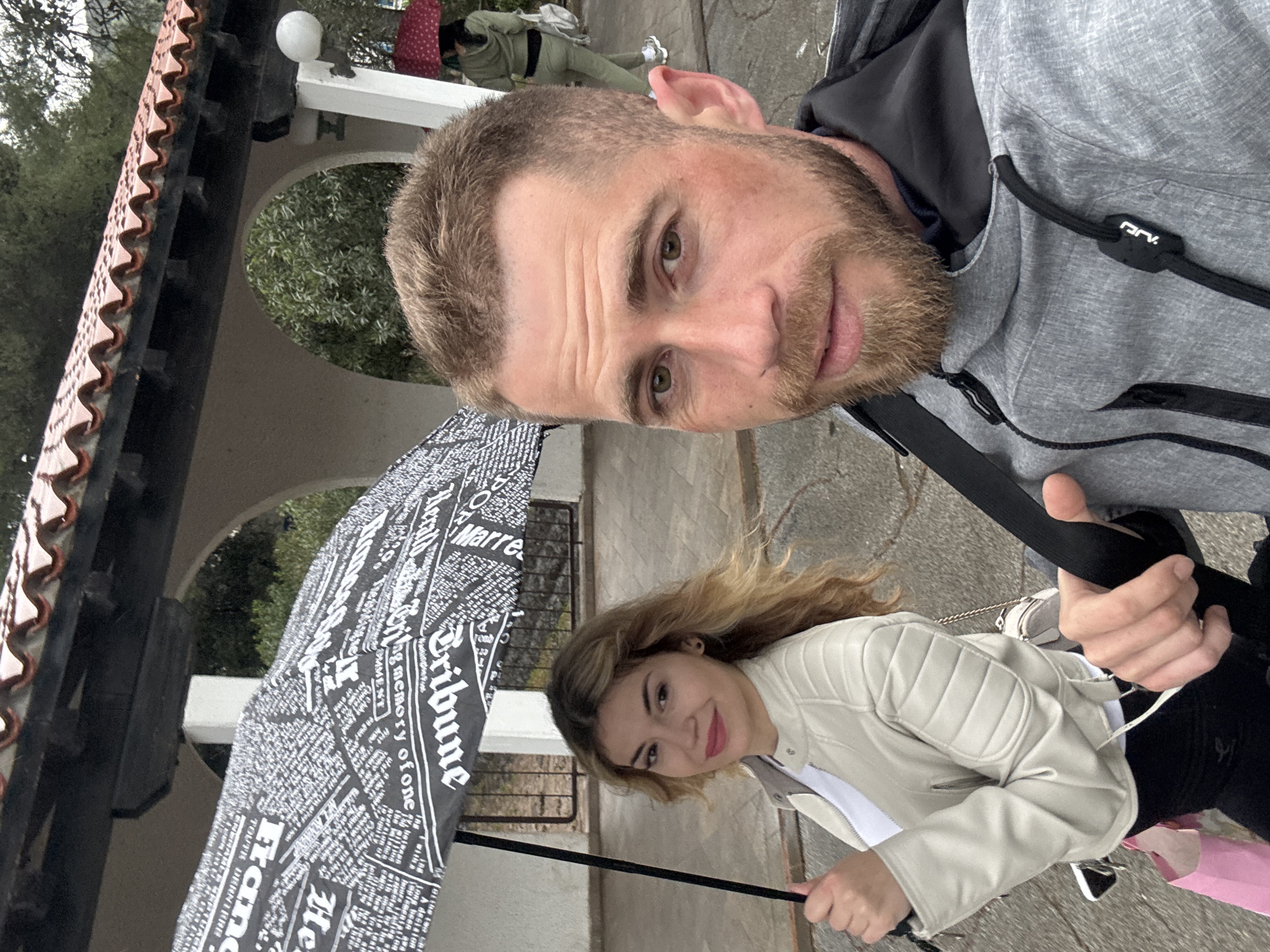 The image is a selfie taken by a man outdoors, with a woman beside him under an umbrella. The man has short brown hair, a beard, and is wearing a gray hooded jacket. The woman has long blonde hair and is wearing a light-colored leather jacket. They are holding a black umbrella with a newspaper print design. The background includes a building with a tiled roof and arched openings. The location is possibly in Budva, Montenegro. Other people can be seen out of focus in the background.