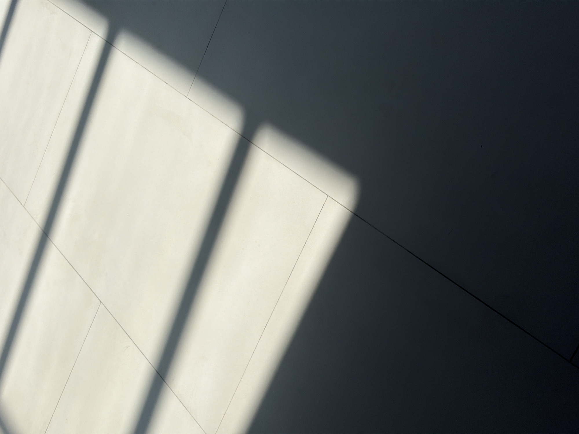 The image shows a section of a tiled floor, partially illuminated by sunlight streaming through a window. The tiles appear to be a light neutral color, possibly off-white or pale gray, with grout lines visible. The sunlight creates distinct bands of light and shadow across the floor. A large portion of the image is in shadow, suggesting a relatively small window or the angle of the sun. The location is unknown. There are no people or other objects visible.