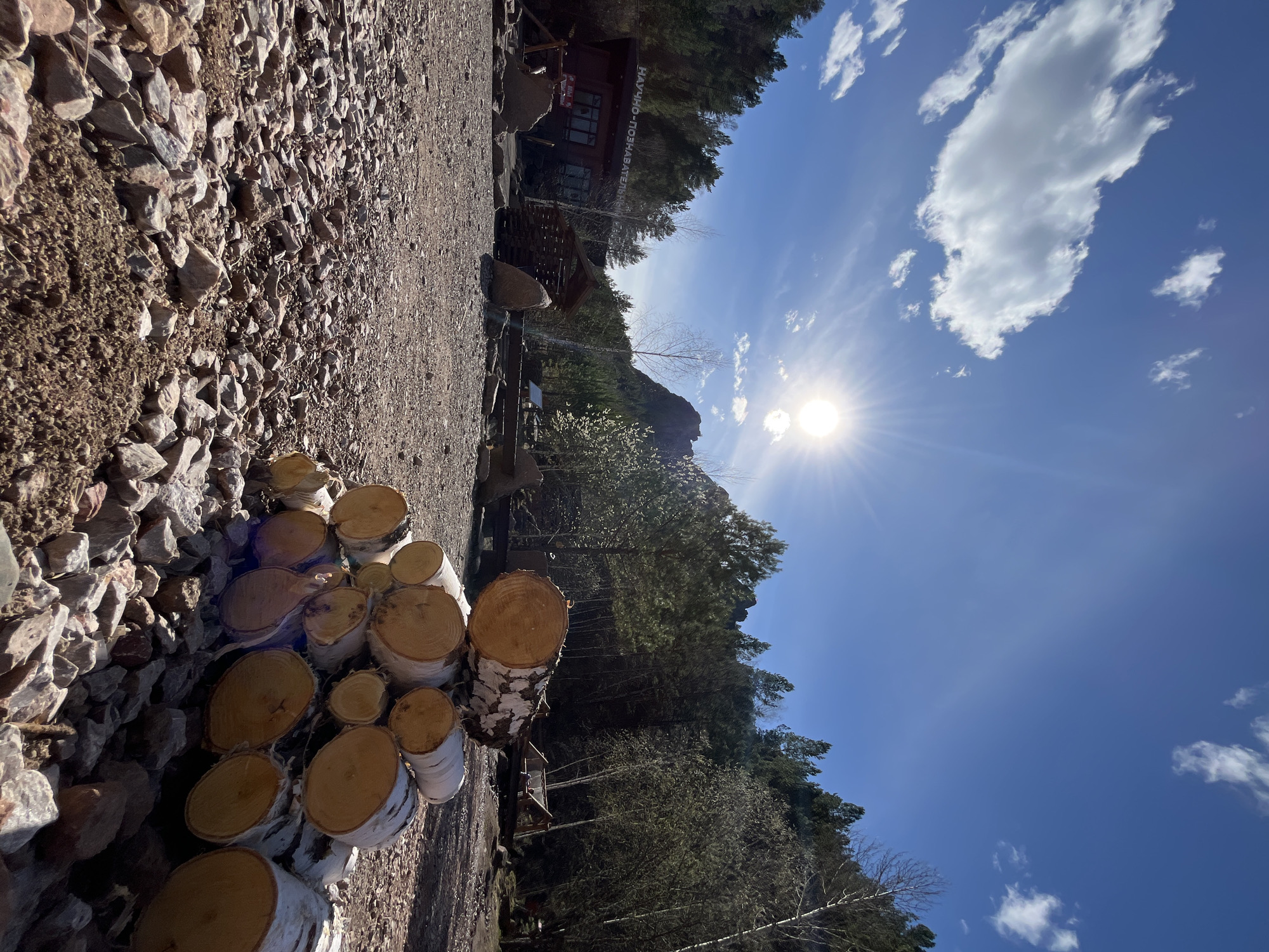 A low-angle, rotated outdoor shot depicts a sunny scene, possibly in the area of Krasnoyarsk, Russia.
In the foreground, a pile of birch logs rests to the left. Next to the logs, there is a field covered with rocks. Behind the logs and rocks is a path leading to an area where there are some picnic tables.
Further back, a building with Cyrillic text can be seen nestled among trees. In the distance, trees and mountains are visible. The sky is blue with a few clouds and a bright sun.