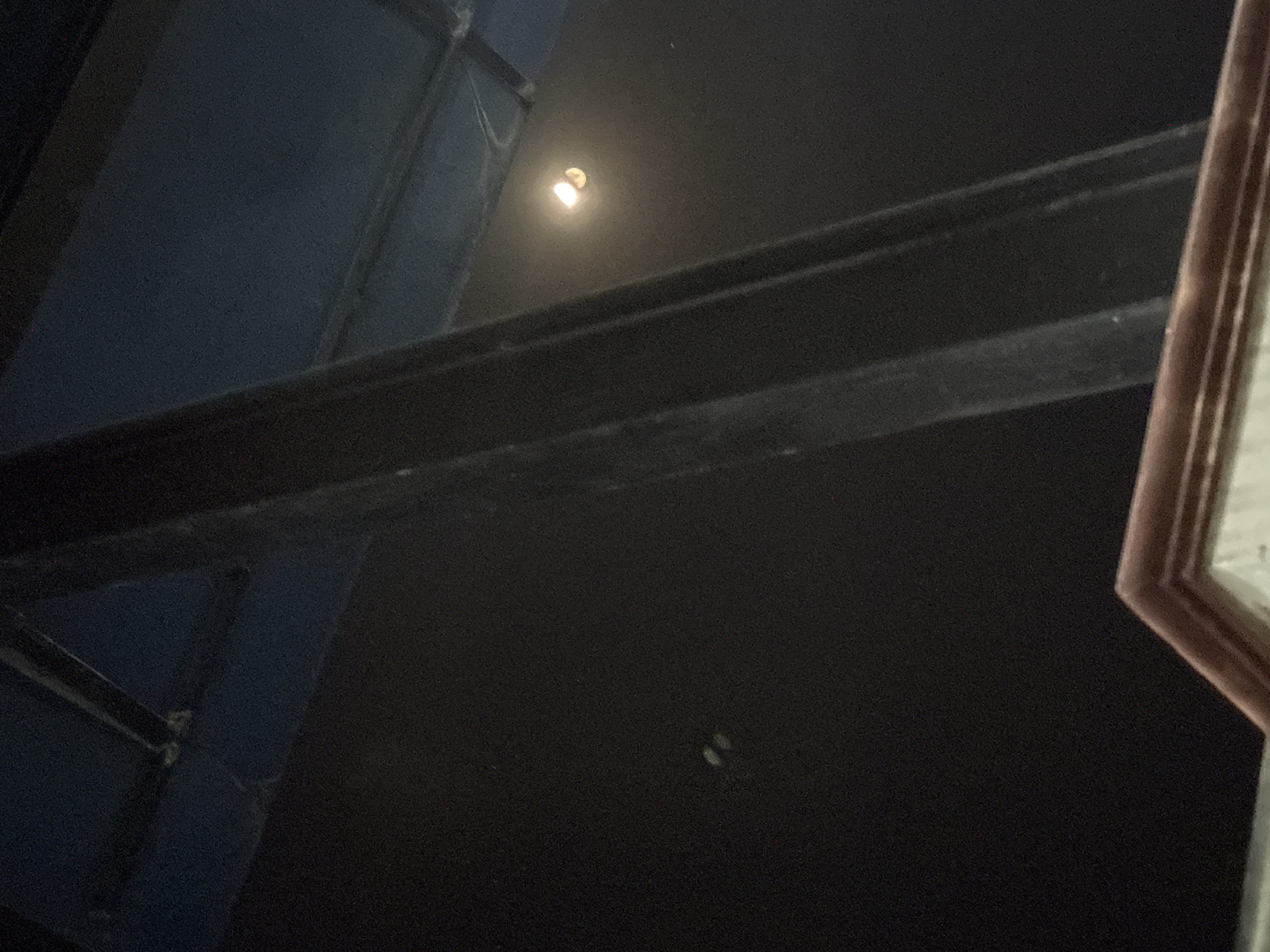 The image appears to be taken inside a dark room, possibly a building or an indoor venue. There is a large, dark, vertical beam in the center of the frame, likely a structural support. Above and behind the beam, a dark blue ceiling or upper wall is visible, with some darker horizontal lines that might be part of the ceiling structure or lighting fixtures.
To the right of the beam, a bright light source with a yellow-orange glow is visible against the dark background. This light appears to be a single bulb or fixture with a reflective element. On the dark wall to the left of the beam, two small, pale, oval-shaped objects are faintly visible.
The bottom of the image shows a portion of a decorative architectural element, possibly a cornice or trim, with a reddish-brown color and layered details.
There are no people or animals clearly visible in the image. The overall impression is of an interior space, likely dimly lit with a focus on the structural elements and a single light source.
Without further information or context, it is impossible to determine the specific location in Gempol, Indonesia. The image itself does not contain any identifiable landmarks or clues that would link it to that particular city or country.