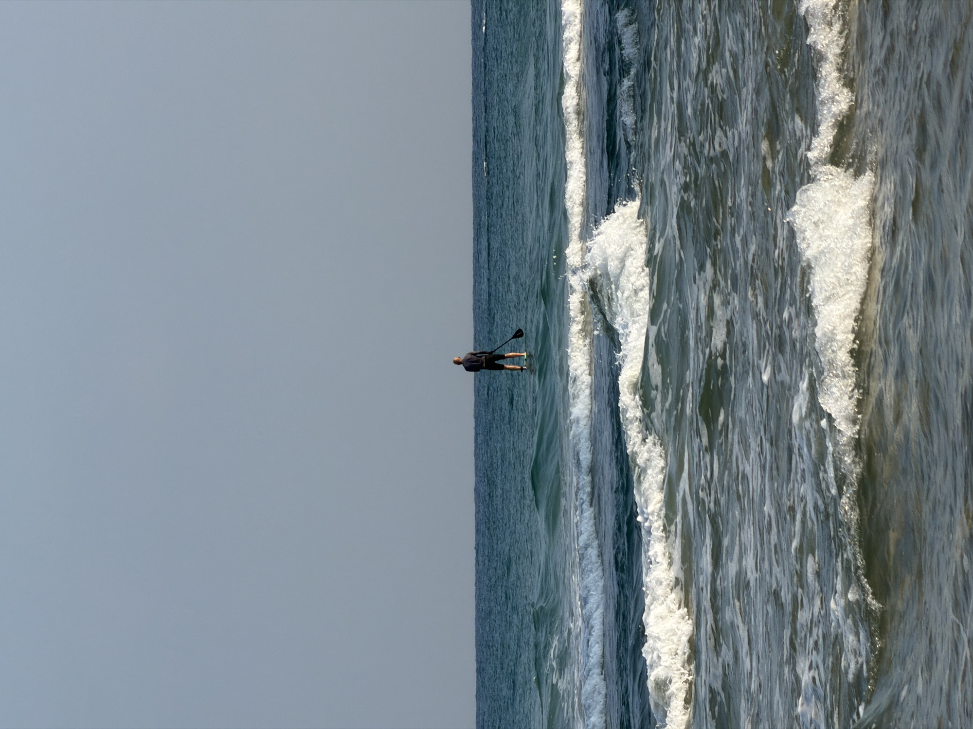 A lone person stands on a paddleboard in the calm, blue waters of the Mediterranean Sea, just off the coast of Bat Yam, Israel. The sky is clear and pale blue, meeting the horizon in a sharp line. The paddleboarder, seen from behind, is wearing a dark shirt and shorts and is holding a paddle. Gentle waves, white-capped and foamy, roll towards the shore in the foreground. The water is a deep, rich blue, with lighter shades where the waves break. The overall impression is one of serene solitude and the vastness of the sea.