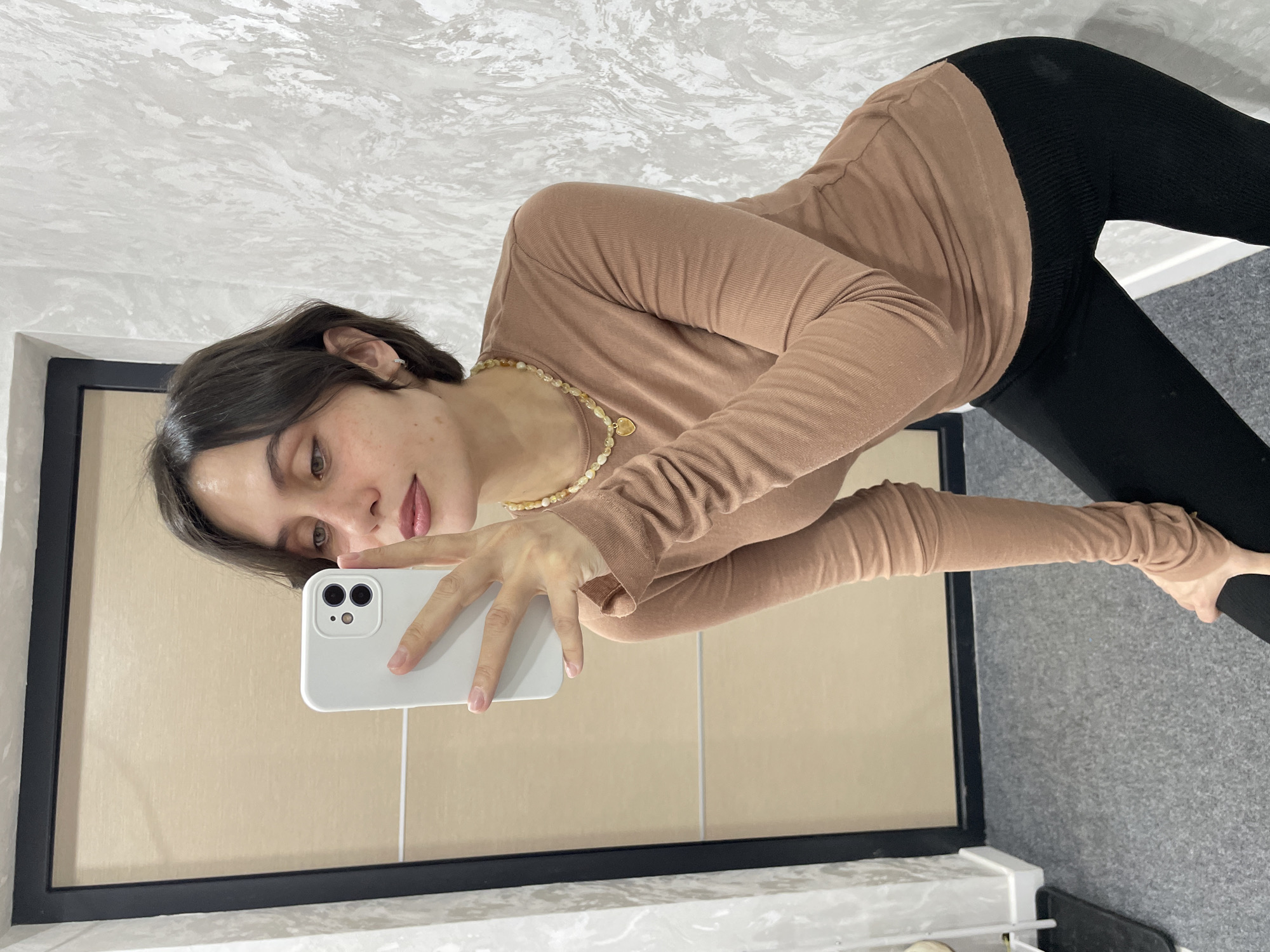 A young woman stands in front of a mirror taking a selfie with a white smartphone. She is wearing a long-sleeved brown top and black leggings. She also has a pearl necklace with a gold pendant. The background consists of a wall with a textured paint finish and a tiled area beneath the mirror. The location is undetermined.
