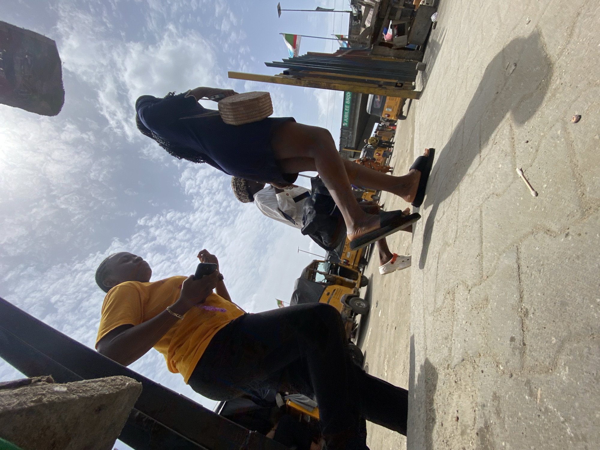 A ground-level, angled shot taken outdoors in Lagos, Nigeria shows three people walking on a concrete surface. The person closest to the camera is wearing a yellow shirt, black pants and holding a phone. Behind and above is a person wearing a navy blue top and short skirt, carrying a woven basket. Further back is another person with a white shirt and dark shorts. The background includes yellow "keke" tricycles, a bridge labeled "JUBILEE BRID", and flags. The sky is visible with patchy clouds.