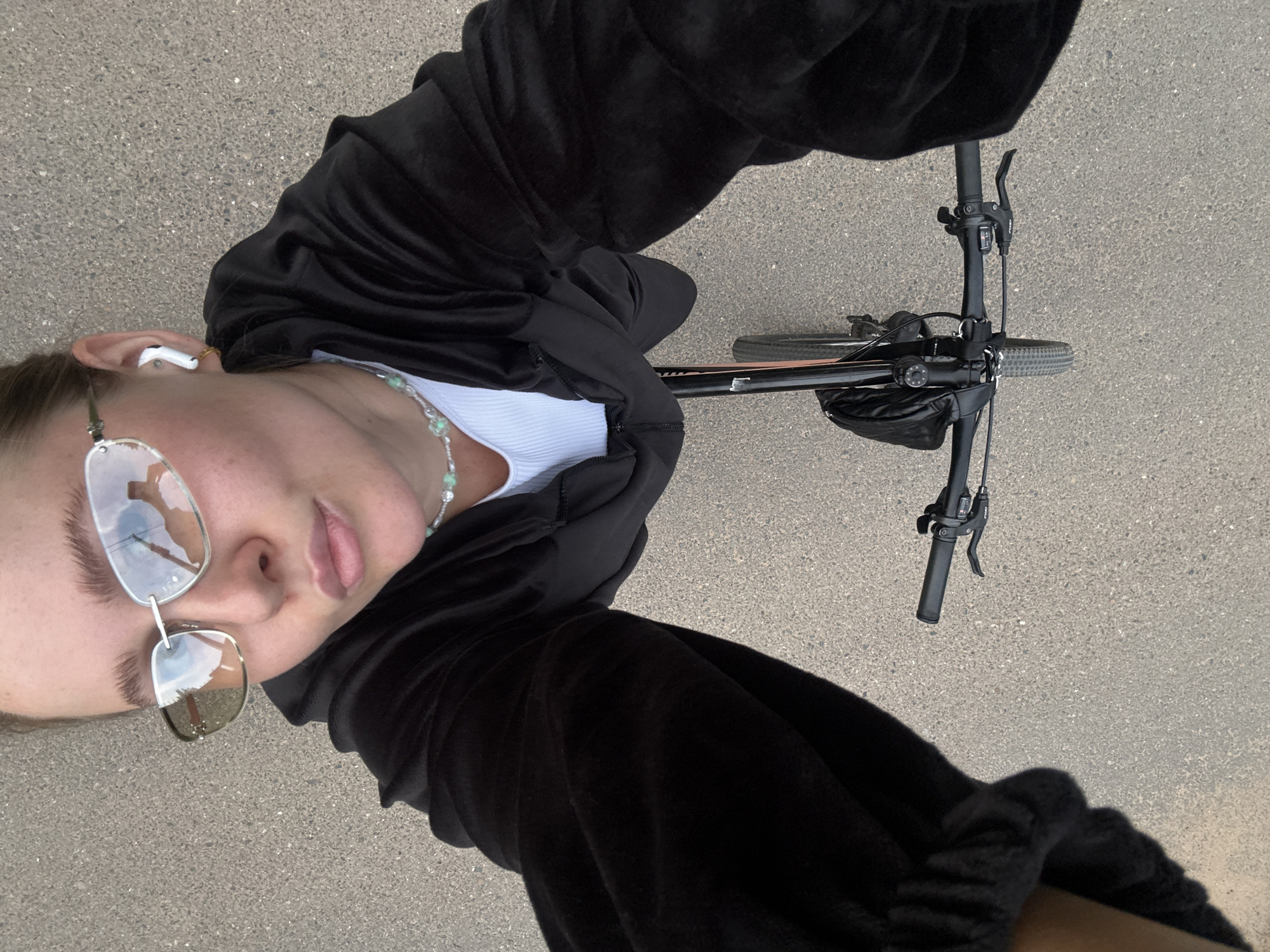 A top-down selfie shows a young woman riding a bicycle on a paved surface, likely a sidewalk or bike path, in Chaikovsky, Russia. She is wearing a black velour tracksuit, a white tank top, a light blue beaded necklace, silver-rimmed sunglasses with tinted lenses, and white wireless earbuds. Her hair is pulled back. The bicycle has black handlebars with silver accents and a black bag attached to the frame. The lighting suggests it is daytime.