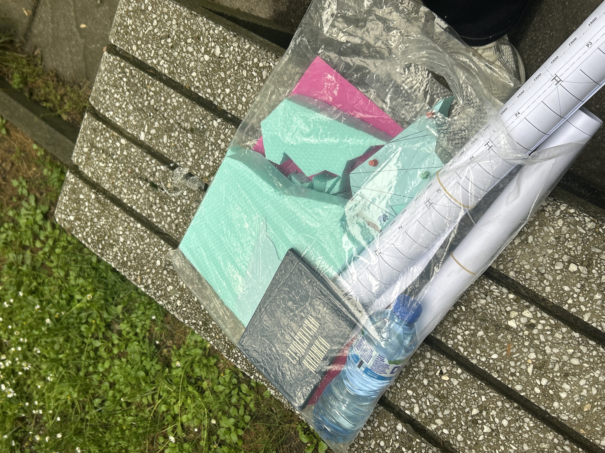 A clear plastic bag filled with various items rests on a grey, speckled concrete bench. To the left and below the bench, green grass with small white flowers is visible. Part of a foot in a white sneaker is seen in the upper background.
Inside the plastic bag, there are several sheets of light green/teal material, along with a pink sheet, possibly craft foam or paper. Two rolled-up white papers, which appear to be technical drawings or blueprints, are also present. These papers feature measurements like "1500" and labels such as "P3," "P4," and "P5," along with symbols resembling "H" and "I." A dark grey or black textured folder or book is included, with visible white text that reads "COMMUNICATION" and a partial word that could be "DESIGN." A plastic water bottle with a blue cap is also inside the bag; the label on the bottle clearly shows "DEVIN," "Вода," and "500 ml."
The scene suggests an outdoor setting during daytime, likely a park or public space, given the presence of the bench and grass. There is no specific activity depicted, but the collection of items could be for an educational project, craft activity, or work-related task.