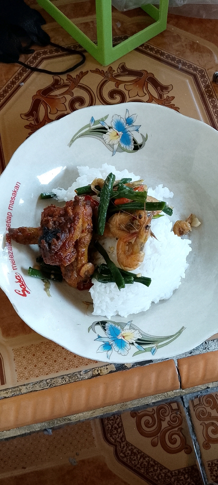A close-up overhead shot shows a white plate with a floral design filled with white rice topped with fried chicken and shrimp stir-fried with green beans and garlic. The plate rests on a table with a patterned brown and tan surface. In the background, a green object, possibly a chair or table leg, is visible. The scene is set in a dining area in Satriamekar, Indonesia.