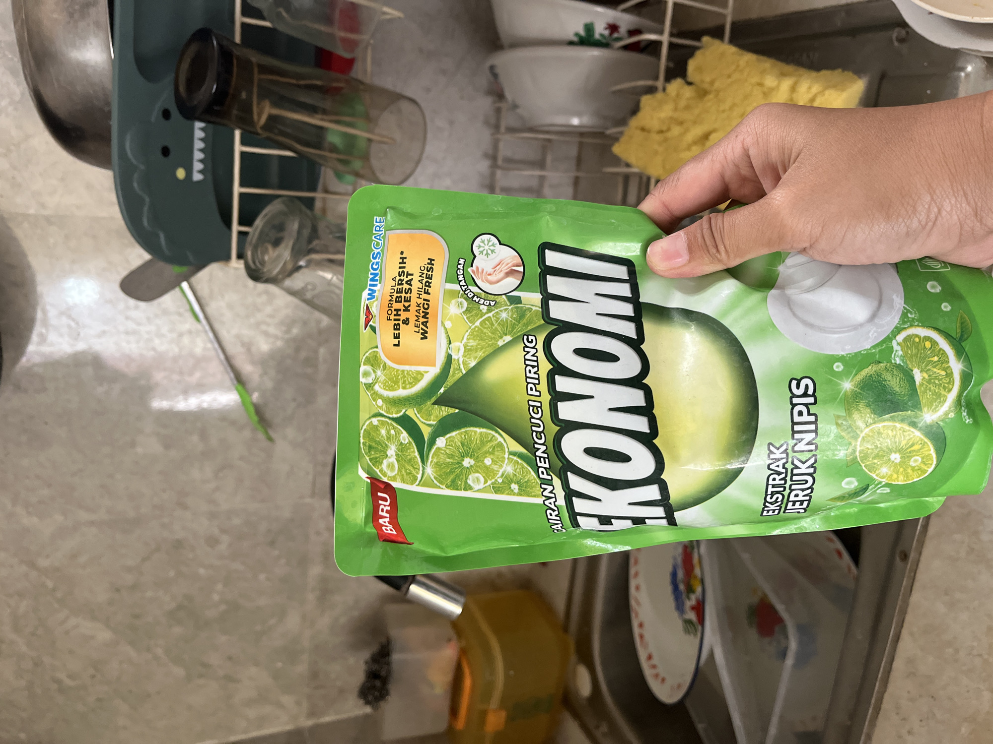 In Kalisampurno, Indonesia, a person's hand is holding a pouch of dish soap labeled "EKONOMI" with "EKSTRAK JERUK NIPIS" (lime extract). The pouch is green and features images of limes and water droplets. The background shows a kitchen sink area with dishes in a drying rack, a sponge, and other kitchen items. The overall scene depicts a common household activity.