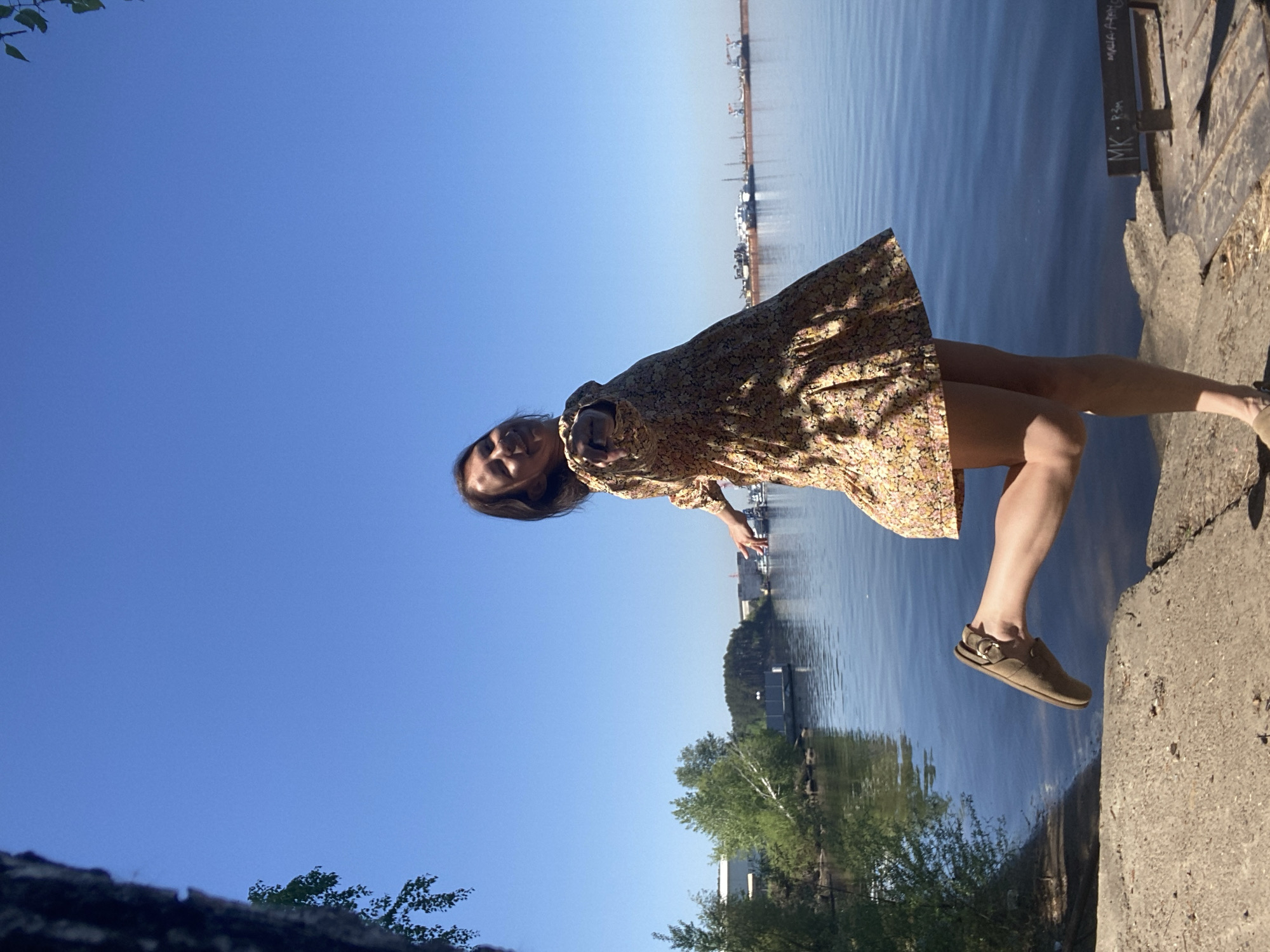 The image shows a woman in a floral dress and tan shoes standing on a concrete embankment next to a body of water. In the background, there are trees, buildings, and industrial structures visible on the opposite shore. The sky is clear and blue. Based on the presence of the concrete embankment and industrial structures on the opposite shore, the scene appears to be on the embankment of Chaikovsky city reservoir, Russia.