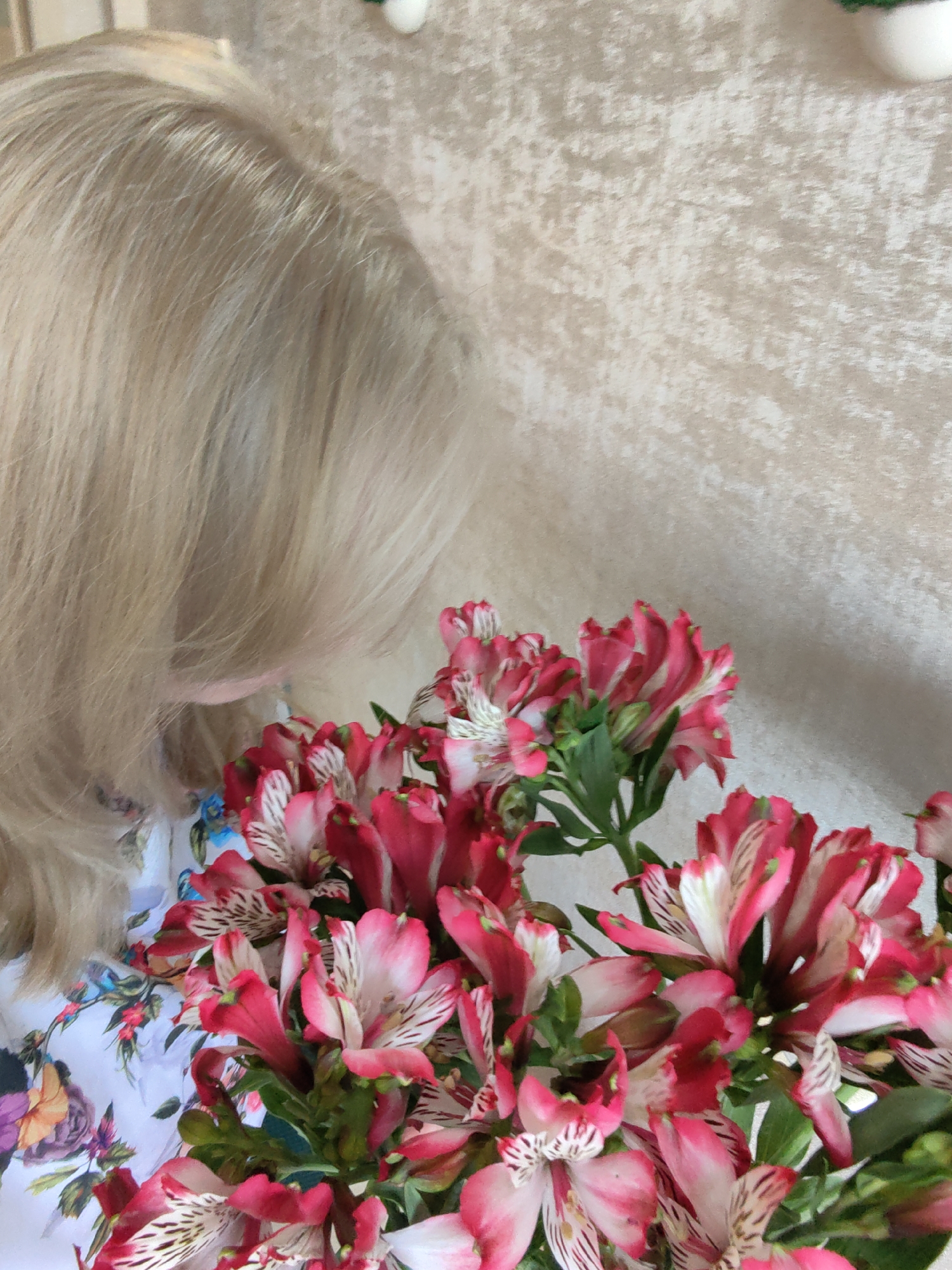 This is a close-up, high-angle, indoor shot of a woman's blonde hair and a bouquet of pink and white alstroemeria flowers. The woman's head is tilted down and to the right, obscuring her face. She is wearing a white garment with a floral print featuring red, blue, and purple flowers. The background is a textured, beige wall. The image appears to be taken in a domestic setting and there is no information to place it in the city of Sochi, Russia.