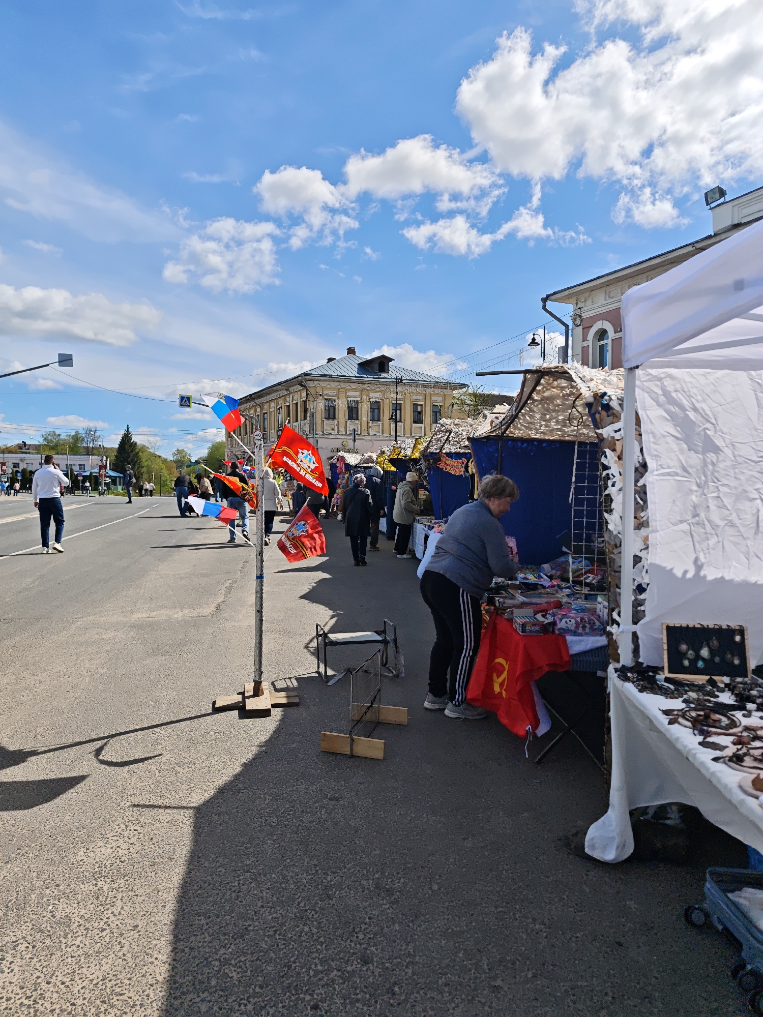 The image captures a street market scene in the city of Uglich, Russia. The foreground features a street with visible asphalt texture. On the right, market stalls are set up under white canopies, displaying merchandise like jewelry and various trinkets. A woman wearing a gray sweater and black-and-white striped pants is examining items on a stall. A red flag with a yellow hammer and sickle emblem is visible on a table. To the left, a metal stand holds red and blue flags. The backdrop shows a street with pedestrians walking along the sidewalk. In the distance, there's a yellowish two-story building with multiple windows. Above, the sky is bright blue with scattered white clouds. A traffic light is visible on the left side of the street, along with additional individuals walking in the distance.