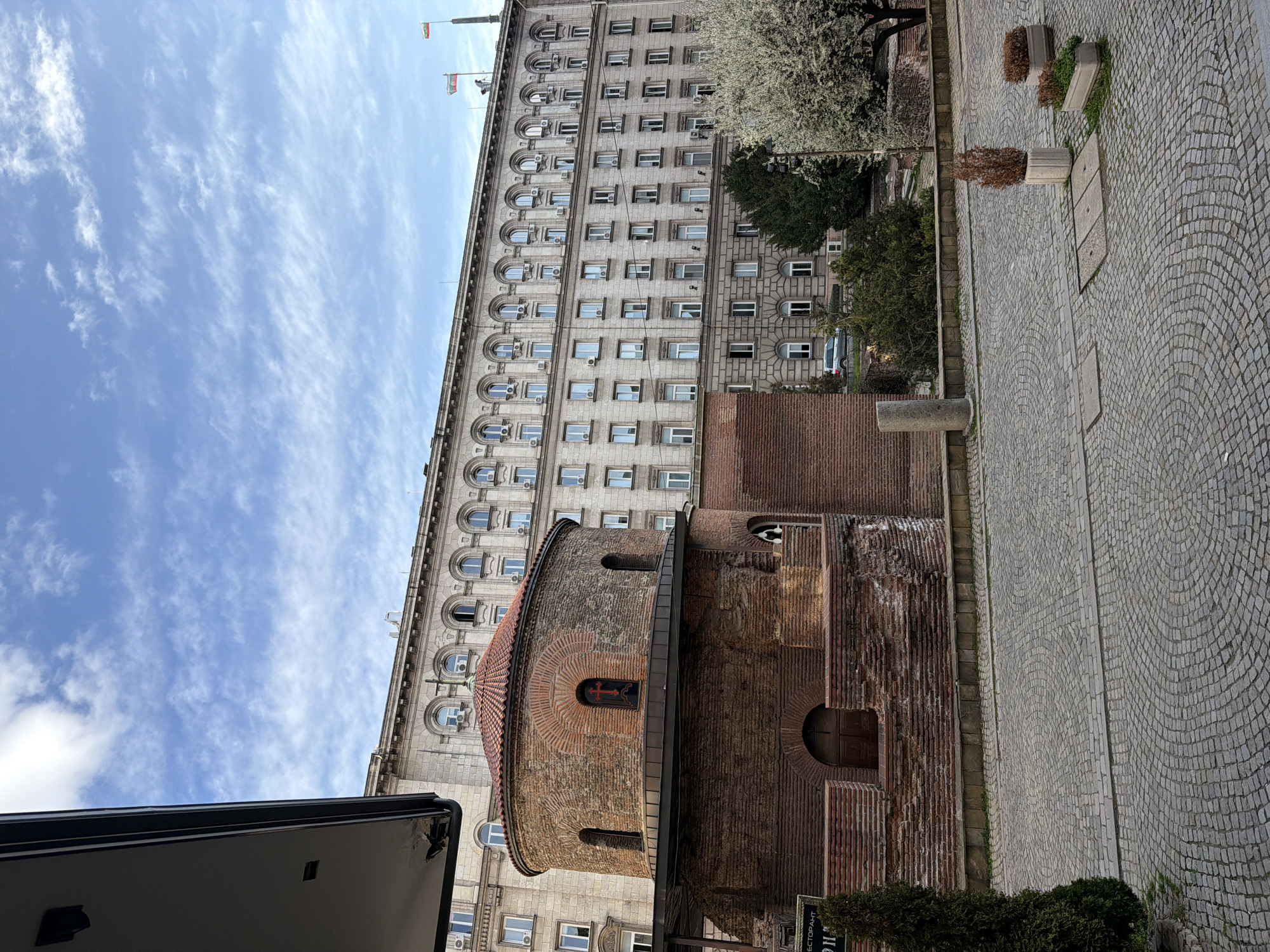 The image depicts the Rotunda of St. George, a historic brick church, situated in the city of Sofia, Bulgaria, against the backdrop of a large, imposing government building. The Rotunda, constructed primarily of reddish-brown brick, features a circular lower section and a smaller, drum-shaped upper section topped with a conical terracotta-tiled roof. Several arched window openings are visible, with one prominent window on the upper level containing a red cross illuminated from within. The building's brickwork exhibits varying textures and patterns, indicating historical construction and potential restoration. A dark, modern-looking canopy or awning partially surrounds the upper portion of the Rotunda, extending from a structure out of frame on the left.
Behind the Rotunda, a multi-story, light-colored stone building with numerous rectangular windows and arched details on its upper floors dominates the background. This building, identifiable as part of the Largo architectural complex (specifically, the Presidency/Council of Ministers building), features a repetitive facade design with multiple exterior air conditioning units mounted below many windows. Two flags, one the Bulgarian national flag and the other possibly the European Union flag, fly from flagpoles on the roofline of this large structure.
The foreground consists of a meticulously laid cobblestone pavement arranged in a fanned, curvilinear pattern. To the left, a tall, slender evergreen tree stands. To the right, a deciduous tree with abundant white blossoms is visible. Two concrete planters containing dried vegetation and one with green sprouts are placed on the cobblestone area. A short stone pillar is also visible near the left edge of the cobblestone. The sky is partly cloudy, with scattered cirrus or altocumulus clouds against a clear blue background. The upper-left corner of the image shows the underside of an overhanging architectural element, possibly an awning or a building edge, with visible dark gutters and some minor wear. No individuals are present in the scene.