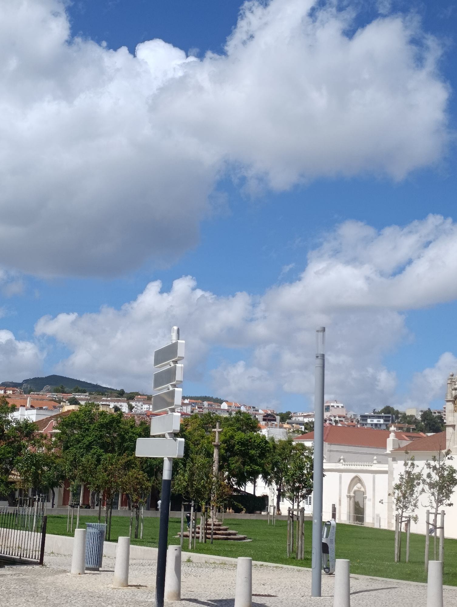 The image captures a street scene in Setúbal, Portugal, on a bright, partly cloudy day. In the foreground, a black pole with a stack of white rectangular signs rises from a cobblestone ground. To the left of the signpost, a silver and blue striped trash can stands between four cylindrical concrete bollards. A metal fence with vertical bars is visible on the far left. Beyond the immediate foreground, a manicured green lawn is dotted with young trees, some with wooden stakes supporting them.
In the middle ground, a white building with a red-tiled roof and arched windows is prominent on the right. To its left, a dense line of mature green trees forms a boundary. Further back, the city of Setúbal sprawls across a hillside, with buildings featuring red-tiled roofs and white facades. A prominent mountain range looms in the distance, under a sky filled with large, fluffy white clouds against a clear blue backdrop. A single street lamp with a clear cylinder stands to the right of the signpost. There are no people clearly visible in the image.