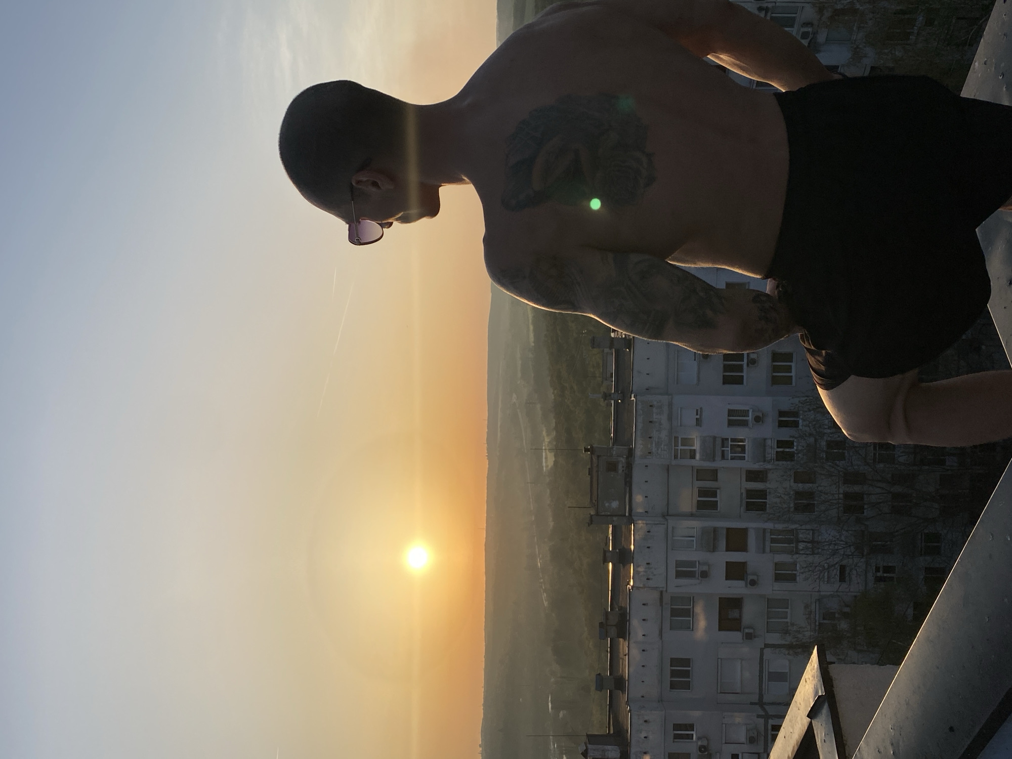 The image captures a shirtless man from the waist up, standing on what appears to be a rooftop or high balcony, gazing towards a vibrant sunset. The man has short hair and pink-tinted sunglasses perched on his head. Visible tattoos adorn his left arm and chest.
The scene is illuminated by the golden light of the setting sun, which is a bright, luminous sphere low on the horizon, creating a warm glow across the sky. The sky transitions from a soft blue at the top to hues of orange and yellow near the sun. A green lens flare is noticeable on the man's chest.
Below the horizon, a hazy landscape of distant fields or hills extends into the distance. In the mid-ground, a large, multi-story concrete apartment building with numerous windows and air conditioning units stands, indicative of an urban or semi-urban environment. The building's architecture suggests an Eastern European setting. The man is positioned beside a dark railing or ledge, indicating a high vantage point. The time of day is clearly sunset, marked by the low sun and warm, directional light. The weather appears clear. No visible text is present in the image. The overall impression is one of peaceful contemplation of the sunset from a high vantage point, consistent with a location like Pleven, Bulgaria.