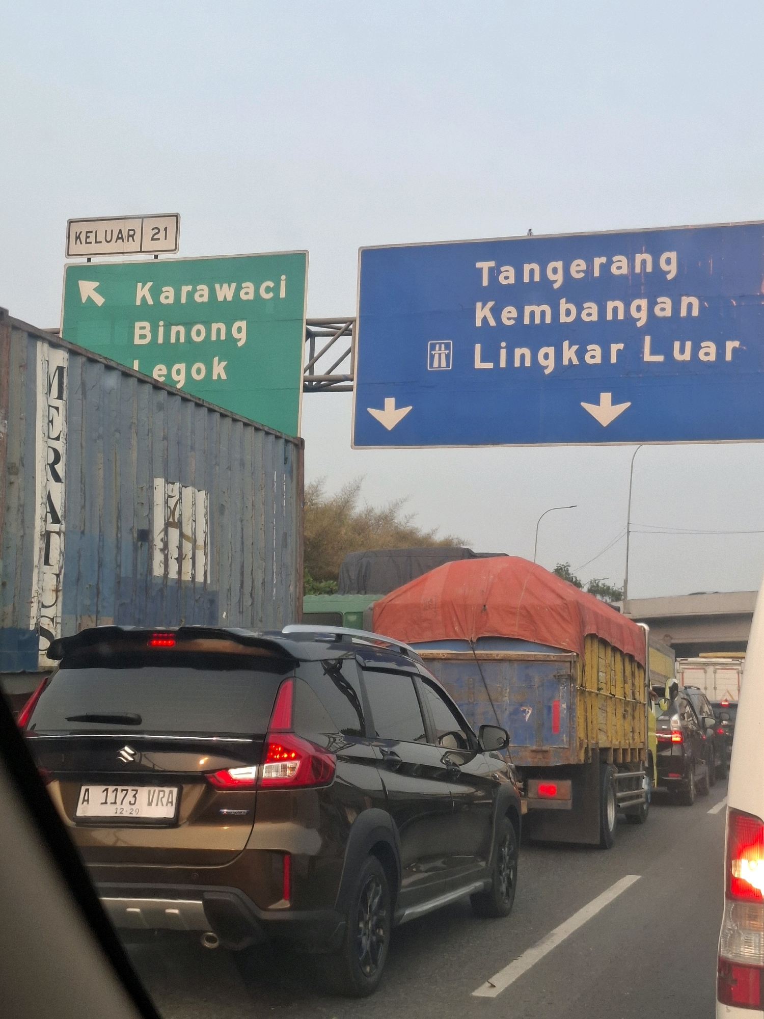 This is a traffic scene in Tangerang, Indonesia. The image features a brown SUV in the foreground with its license plate visible, showing "A 1173 VRA". Behind it, there is a truck with a red tarp covering its load. Further ahead, other vehicles are seen in traffic. Overhead signs indicate directions: one green sign with a left arrow points to "Karawaci", "Binong", and "Legok", and a blue sign directs towards "Tangerang", "Kembangan", and "Lingkar Luar" with downward arrows. The environment appears to be a highway or major road with multiple lanes. The sky is clear and overcast.