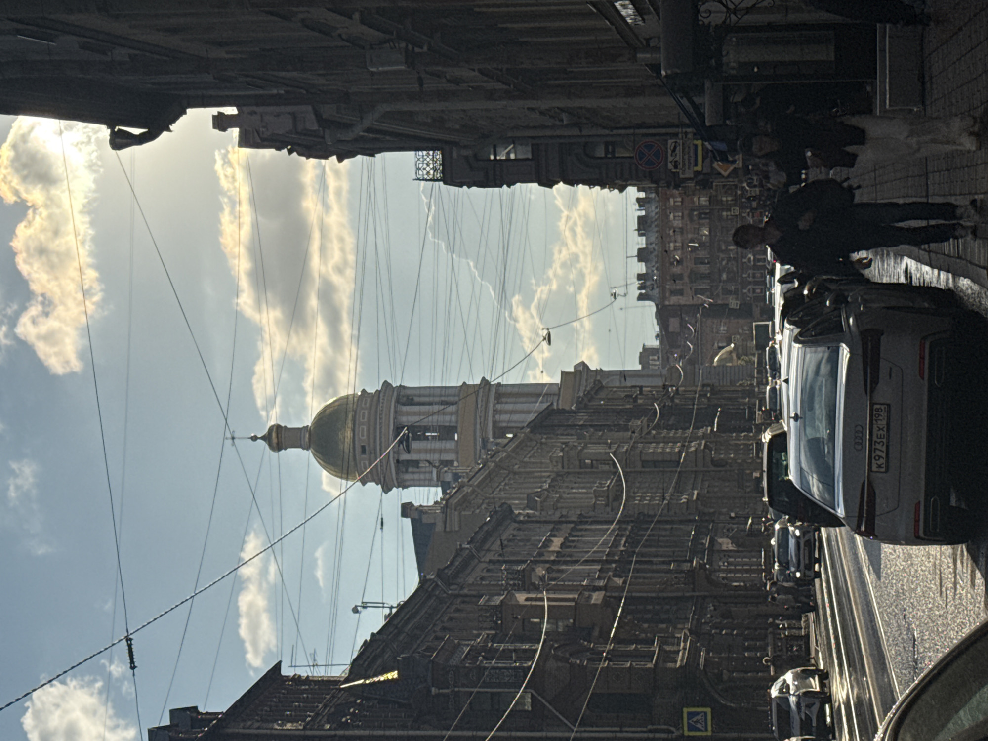 A street scene in St. Petersburg, Russia, features a golden-domed church tower and classic European architecture. The street is wet, suggesting recent rain, and several cars are visible, including an Audi in the foreground. People walk along the sidewalk, some with bags. The sky is partly cloudy with bright sunlight. Overhead, a network of wires crisscrosses the scene. The overall impression is of a bustling urban environment with historical influences.