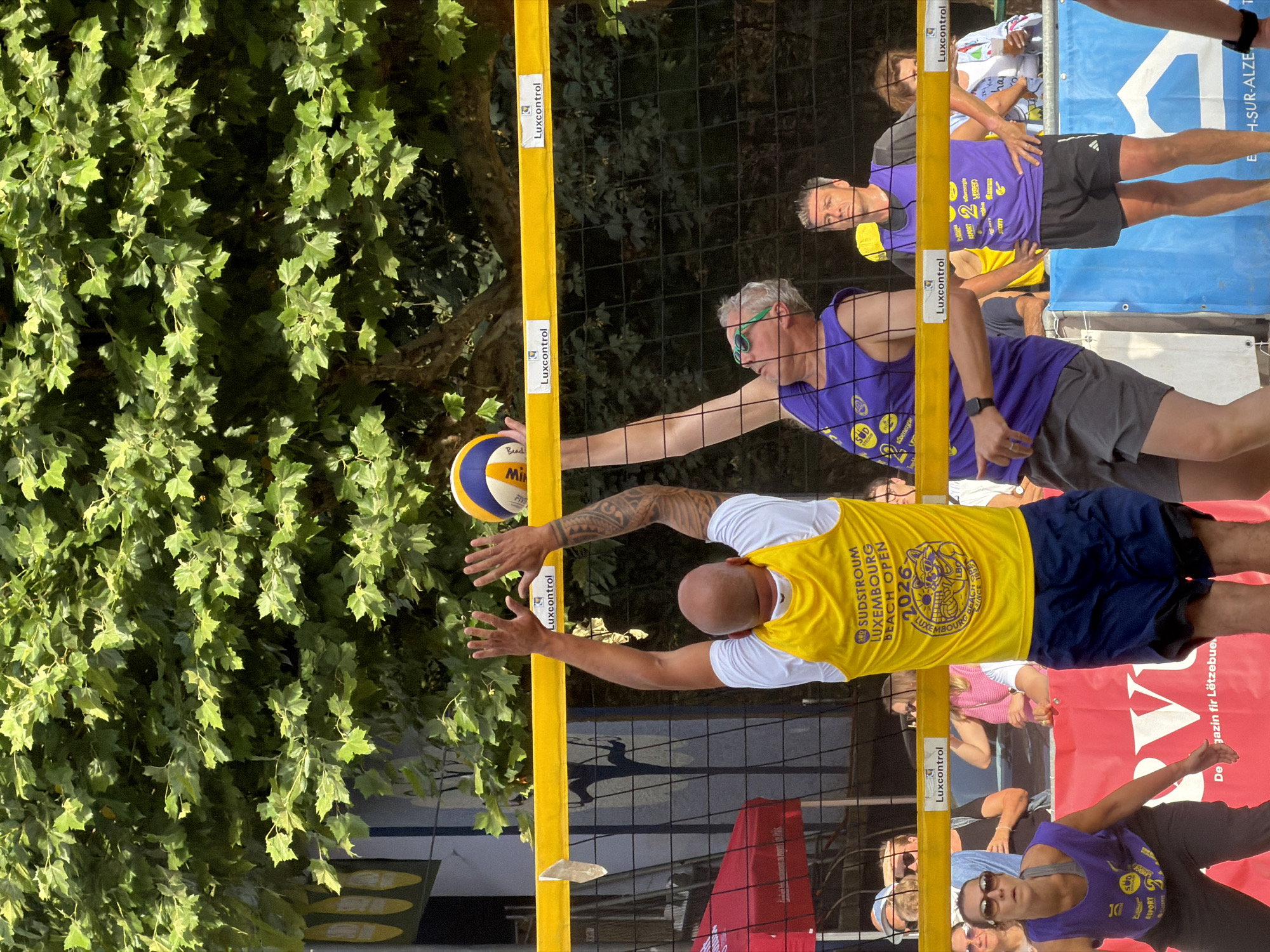 A beach volleyball match is underway on a sunny day under a canopy of green trees. In the foreground, two male players are engaged in a play near the net. One player, wearing a yellow tank top with "SÜDSTRÖM LUXEMBOURG BEACH OPEN 2026" printed on it, is in the process of spiking a volleyball. He is bald and has tattoos on his left arm. His opponent, wearing a purple tank top and green sunglasses, is reaching up, possibly to block the spike. Behind them, other players and spectators are visible. The volleyball court has a yellow net with "Luxcontrol" branding. The backdrop suggests an outdoor venue in Esch-sur-Alzette, Luxembourg, with trees and buildings visible beyond the court. A red tent is set up to the left of the court, and a blue banner with white lettering is visible at the bottom of the image.