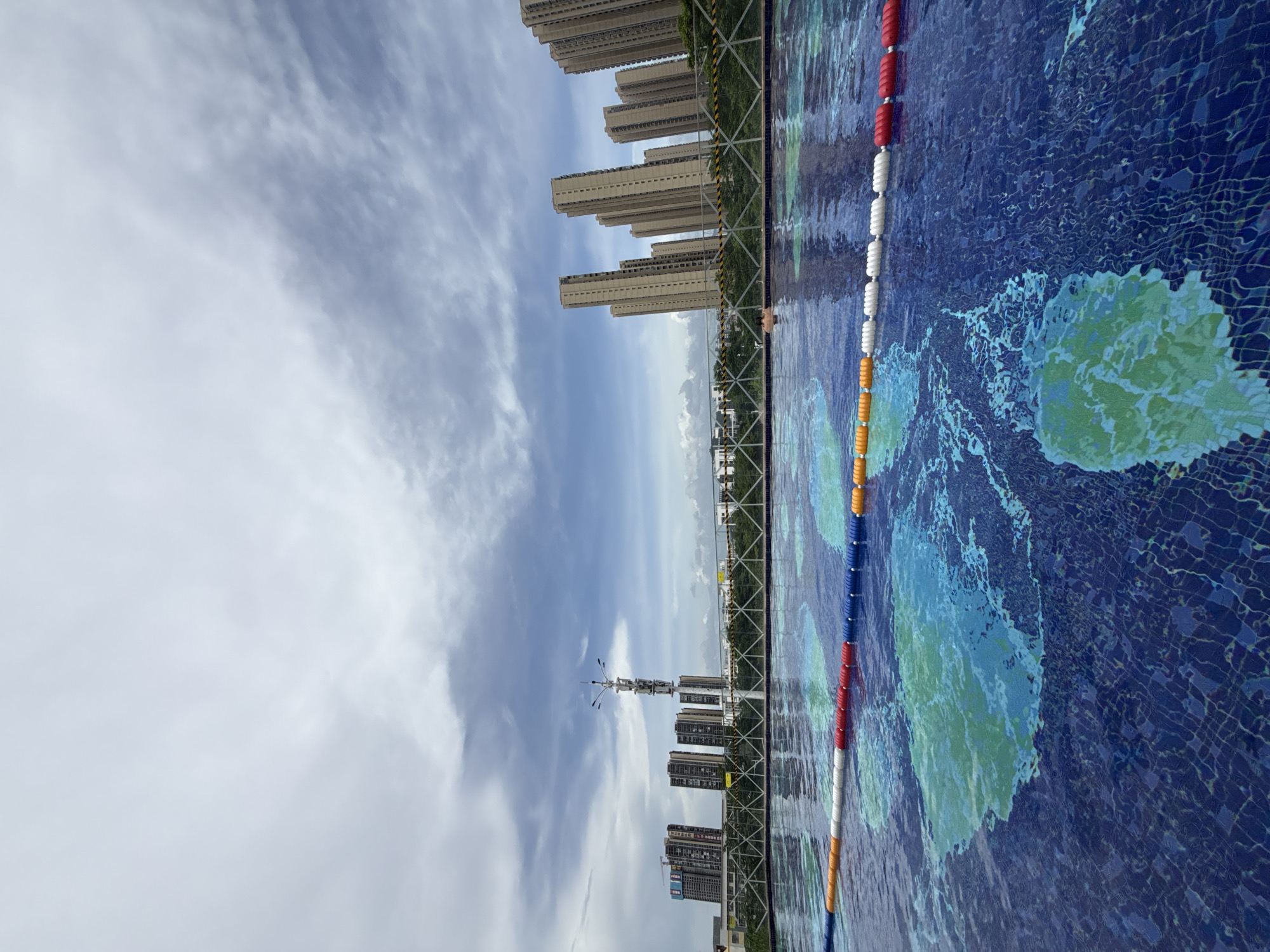 This image depicts a rooftop swimming pool with a view of the city of Huizhou, China. The pool is tiled with a blue mosaic pattern that resembles a map of the world. Lane ropes made of red, white, and blue buoys divide the pool. In the background, several tall residential buildings are visible, along with a distinctive telecommunications tower. The sky is cloudy with patches of blue. A person is partially submerged in the water on the right side of the frame. A metal fence with yellow and black hazard tape runs along the edge of the pool area.