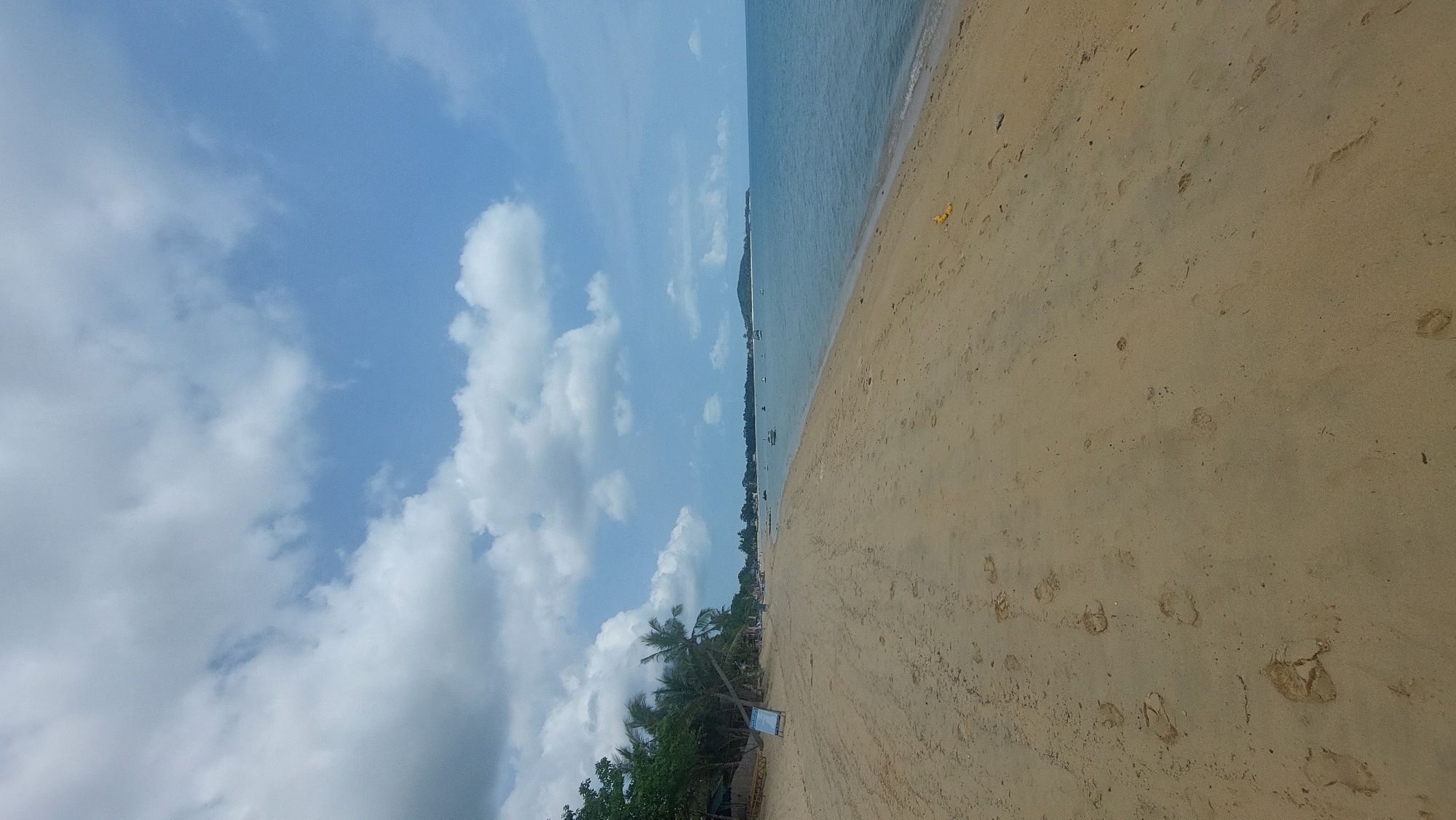 The image displays a serene tropical beach scene, likely captured during daytime under bright, sunny weather with scattered white cumulus clouds in a clear blue sky. The location context indicates Baan Mae Nam, Thailand.
The scene features a wide expanse of light brown sand in the foreground and extending into the mid-ground. Numerous footprints are visible in the sand, suggesting recent human activity. A few small, indistinct pieces of debris, including some yellow items, are scattered on the beach.
To the left of the sandy beach, a strip of lush green tropical vegetation is visible, predominantly featuring palm trees whose fronds reach upwards. A white sign with blue lettering is partially visible near the base of the vegetation, but the text is too small to be deciphered.
The calm, light blue ocean stretches from the shoreline into the distance, appearing tranquil. Several small boats, possibly longtail boats or fishing vessels, are dotted across the water near the coast, fading into the horizon. A distant landmass or hill is discernible across the water, forming the far horizon line.
The image conveys a peaceful and idyllic atmosphere, characteristic of a tropical coastal environment. No individuals are directly visible in the frame, but the footprints imply human presence on the beach.