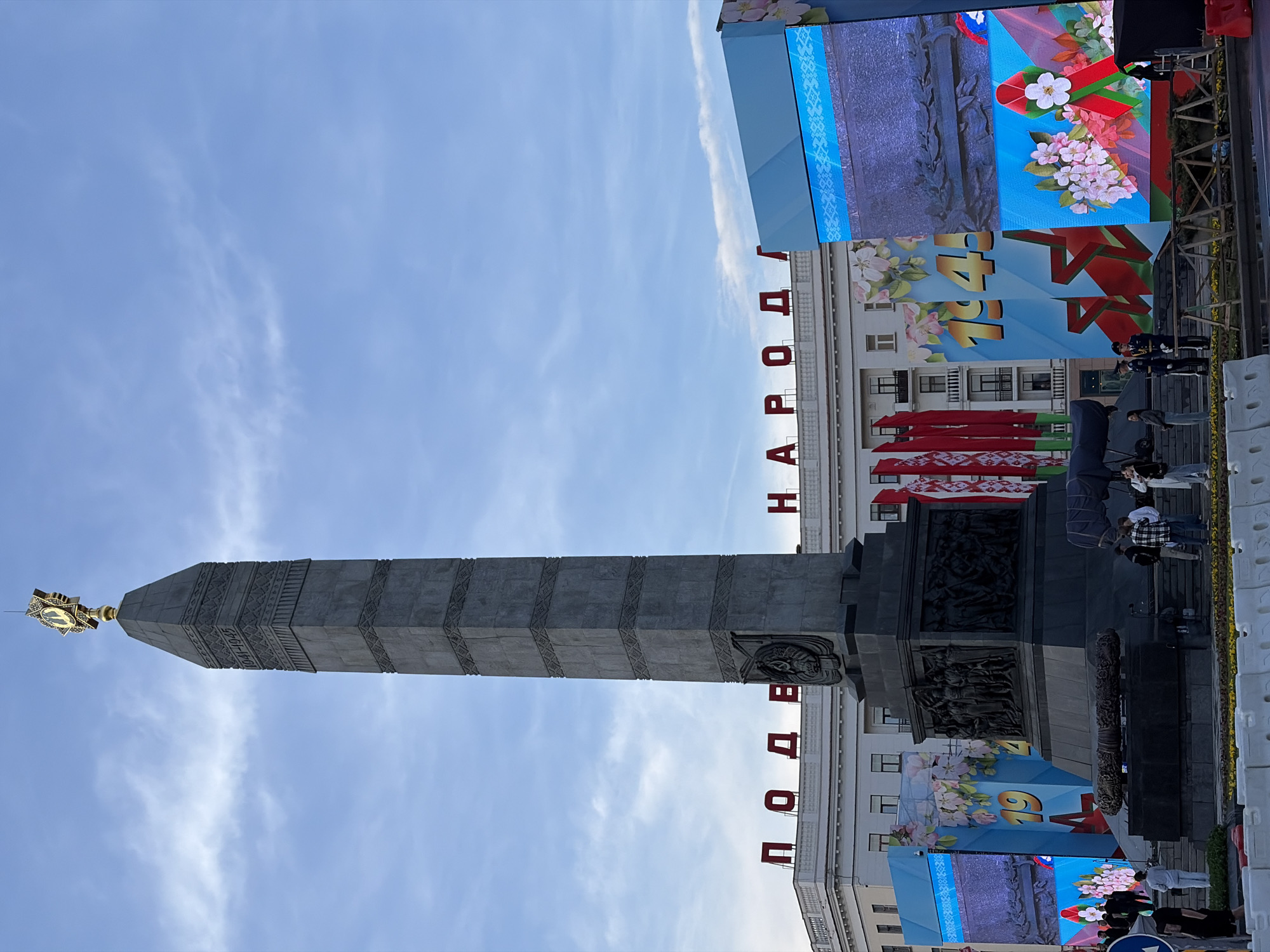 The image shows the Victory Monument in Minsk, Belarus, viewed from a low angle. The monument is a tall, grey, obelisk-shaped structure with decorative elements. At its base, there are reliefs depicting figures and scenes. At the top is a golden star. Behind the monument is a light-colored building with lettering in red on its roof. The letters spell out "ПОДВИГ НАРОДА" and mean "Feat of the people". Blue banners or screens are positioned around the monument and building, decorated with floral patterns, the year 1945, and red ribbons. Several people are standing and walking near the monument. The sky is light blue with wispy clouds.