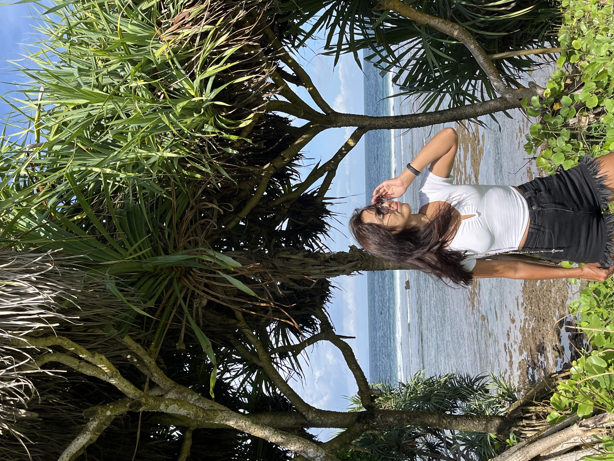 A woman stands on a beach in Kuta Selatan, Indonesia, on a sunny day. She is wearing a white short-sleeved shirt, black shorts, and sunglasses. She is also wearing a dark colored wristband on her left wrist. She has long, dark hair. She is standing in front of a variety of trees and bushes. The beach is visible in the background. The ocean is a light blue color.