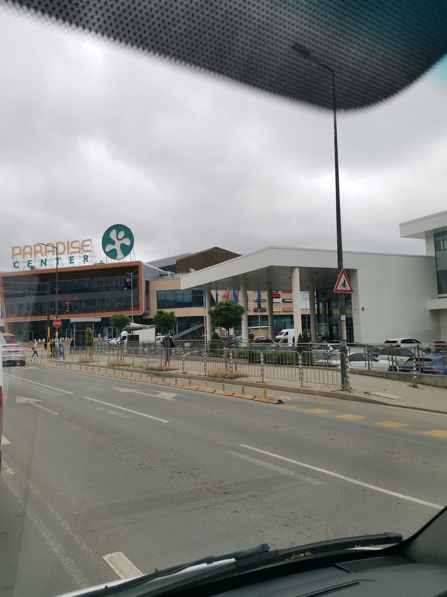 A street view shows the Paradise Center mall in Sofia, Bulgaria. The mall has large windows and is decorated with a green circular logo. There are several cars on the road, and some people are walking on the sidewalk. The sky is overcast, and the overall scene is somewhat muted. A streetlamp with a triangular sign depicting a bicycle is visible.