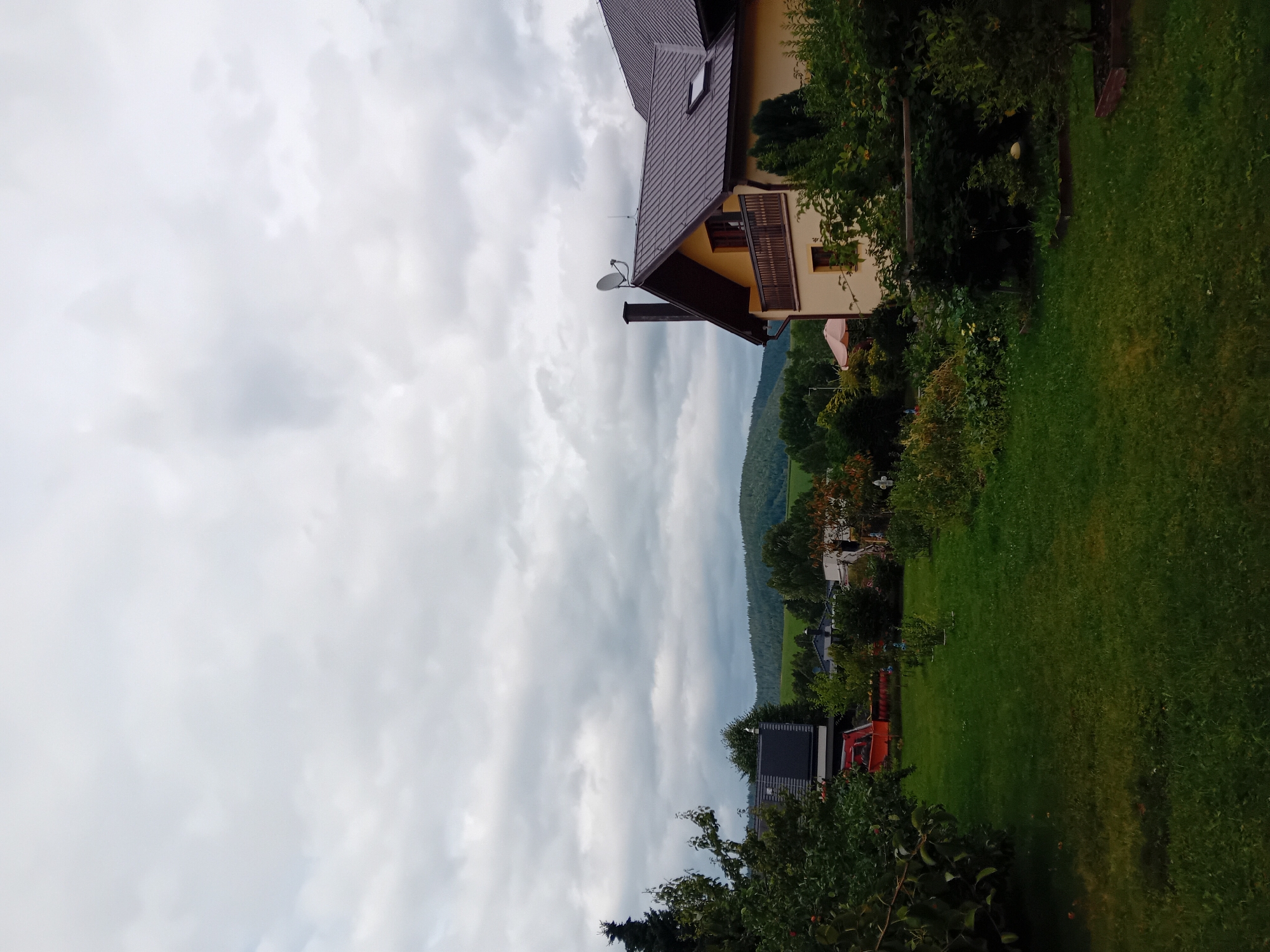 The image depicts a residential property in Zuberec, Slovakia, under an overcast sky. In the foreground, a well-maintained green lawn stretches towards the viewer, with various garden plants and trees bordering its edges. To the right, a section of a two-story house is visible, characterized by its cream-colored facade, brown roof, and a wooden balcony. A chimney with a satellite dish is prominently featured on the roof. To the left, another building with a dark-tiled roof and solar panels is partially seen, adjacent to a red vehicle. In the midground, dense green foliage and trees create a natural buffer, obscuring other structures. The background reveals rolling hills covered in lush green forests, characteristic of the Slovak landscape. The overall scene suggests a peaceful, rural setting within the town of Zuberec.