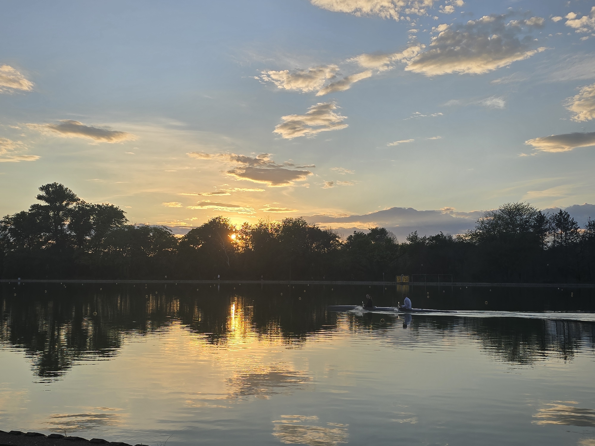 The image captures a tranquil sunset scene over a body of water, likely a lake or large pond. The sun is positioned low in the sky, partially obscured by trees along the shoreline, casting a warm golden glow across the water and sky. Clouds are scattered across the blue sky, with varying shades of orange and pink from the sunset. The water reflects the colors of the sky and the silhouette of the trees, creating a mirrored effect. Two figures are visible in a rowboat on the water, engaged in rowing. The location appears to be a park or recreational area, as indicated by the natural environment and the presence of rowers. There is a sign or marker near the shoreline with numbers "750 750" on it. The city and state are unknown.