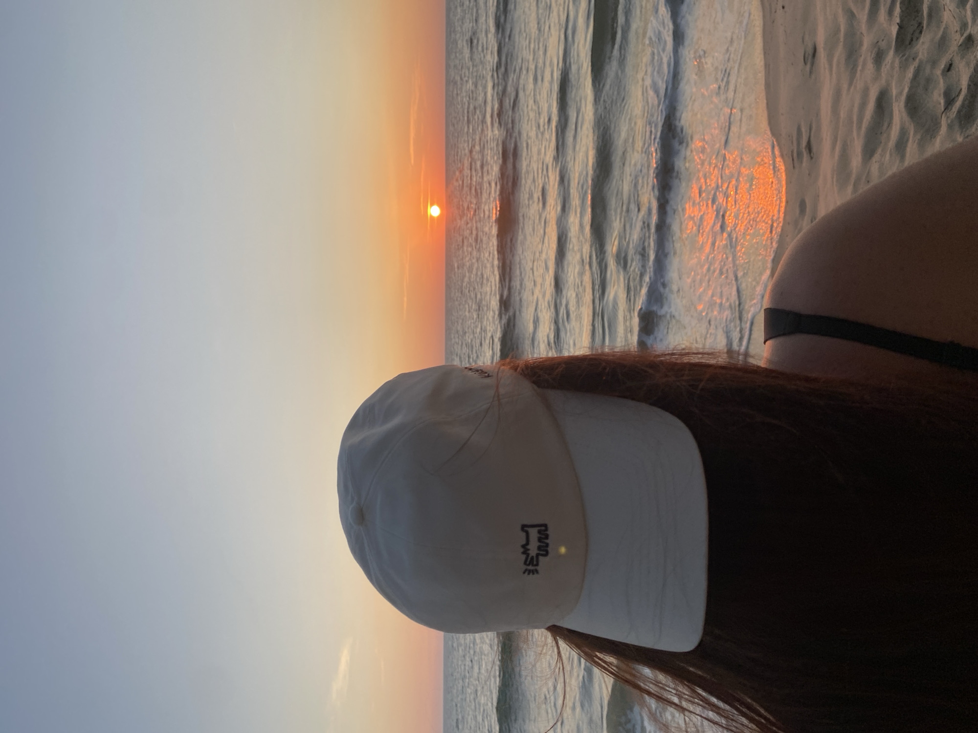 A person wearing a white baseball cap is standing on a beach, with red-brown hair visible beneath the cap. The cap has a small, dark-colored design embroidered on the side. The person is facing the ocean during sunset. The sun is a bright orange disc near the horizon, and its reflection is visible on the water's surface. Waves are washing onto the sandy beach. The location is Choeng Thale, Thailand.